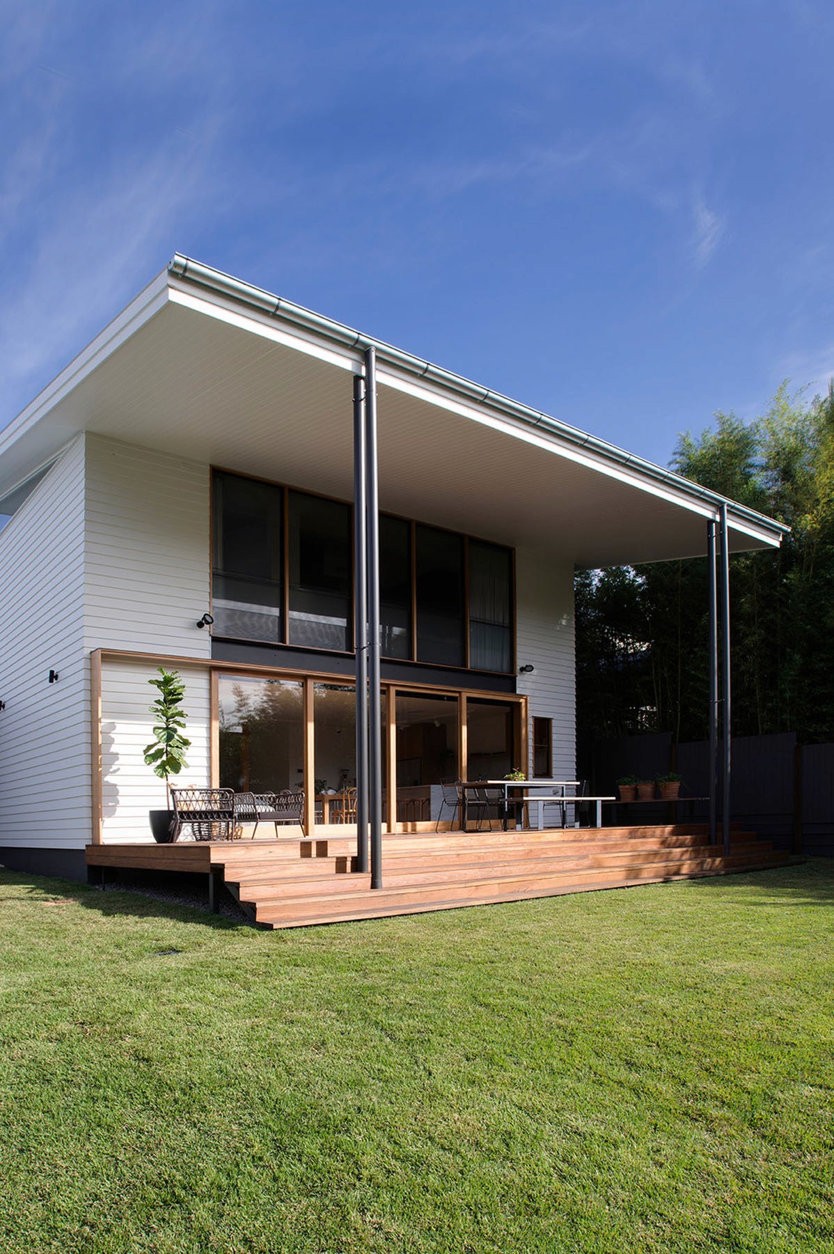 Covered outdoor deck with exposed structure, slender steel posts, and full-width timber steps opening to the lawn.