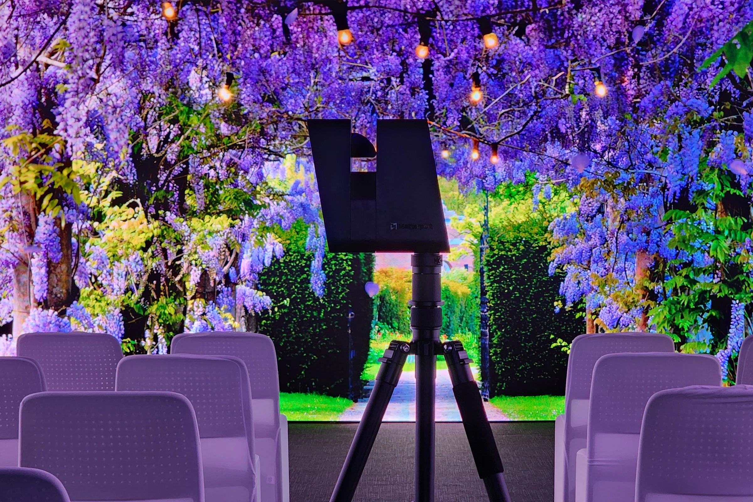 A Amtterport Pro3 professional 3D spatial data camera mounted on a tripod, positioned in front of white event seating and a vibrant purple wisteria light projection at ILA Immersive Light and Art. Captured by ABM visual.