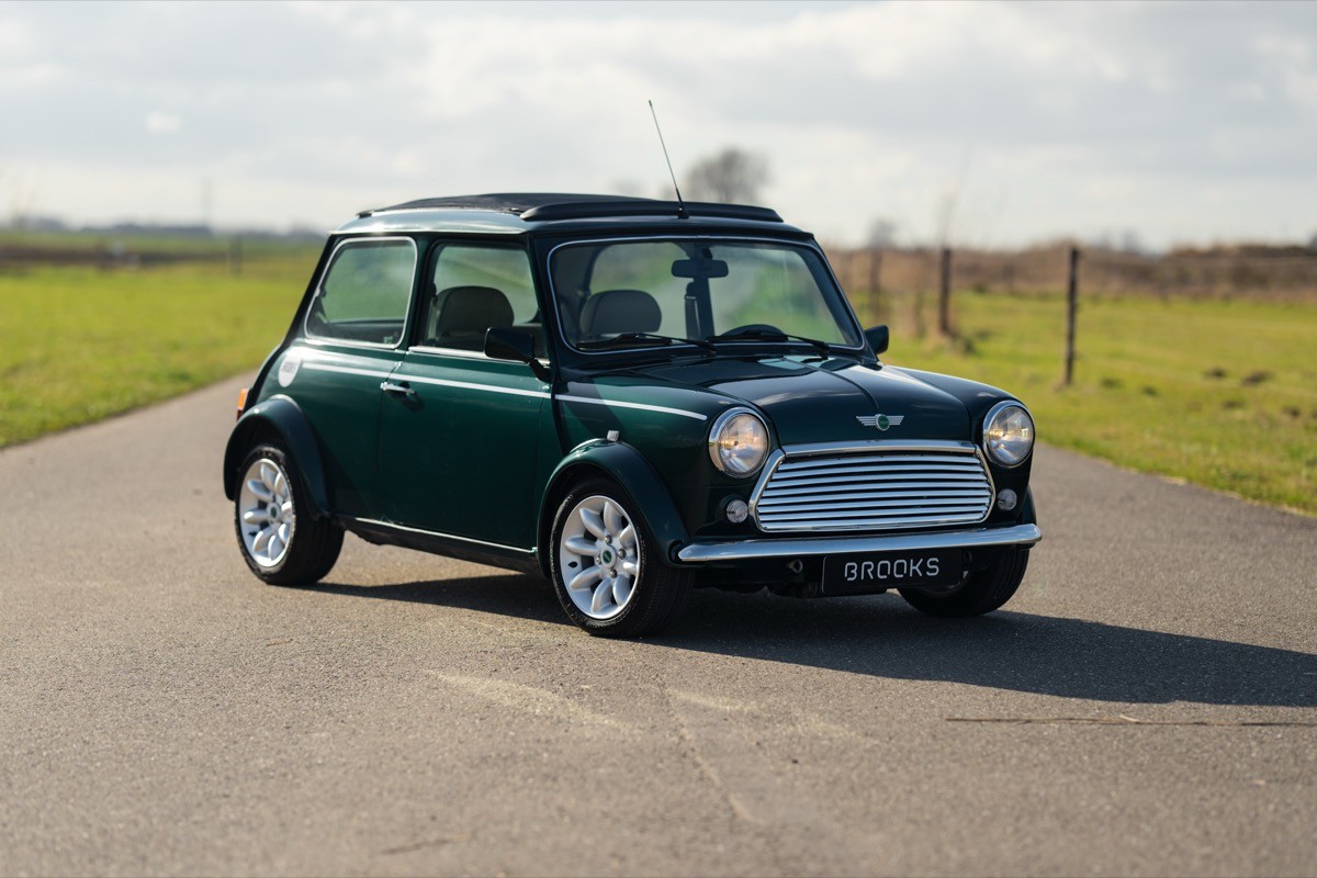 A classic green car parked on a paved road with grass fields in the background under a cloudy sky.