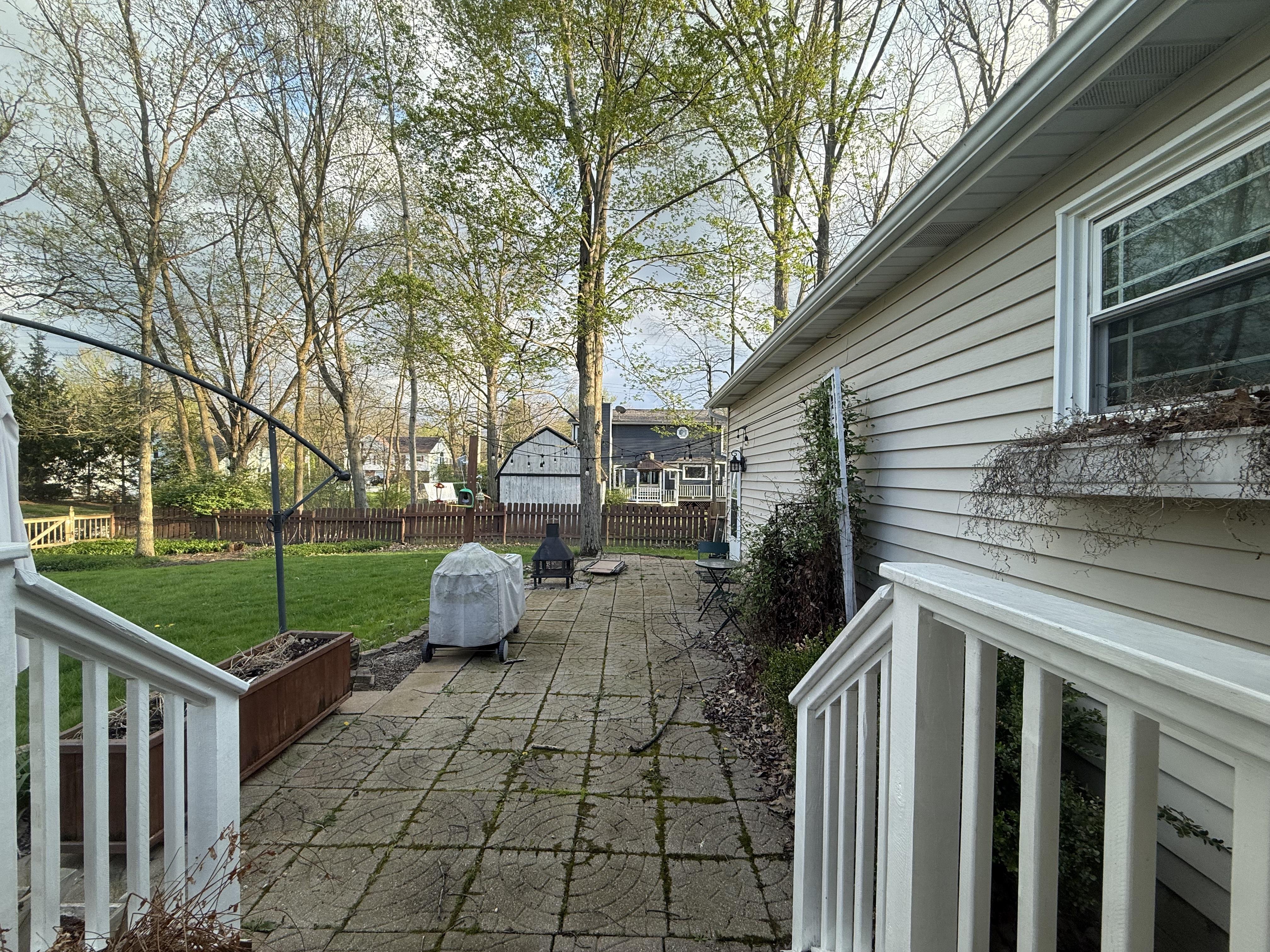 The original patio. Moss-covered square pavers, a rusty raised garden bed on the left, a covered grill, a small fire pit at the far end, white vinyl siding house
