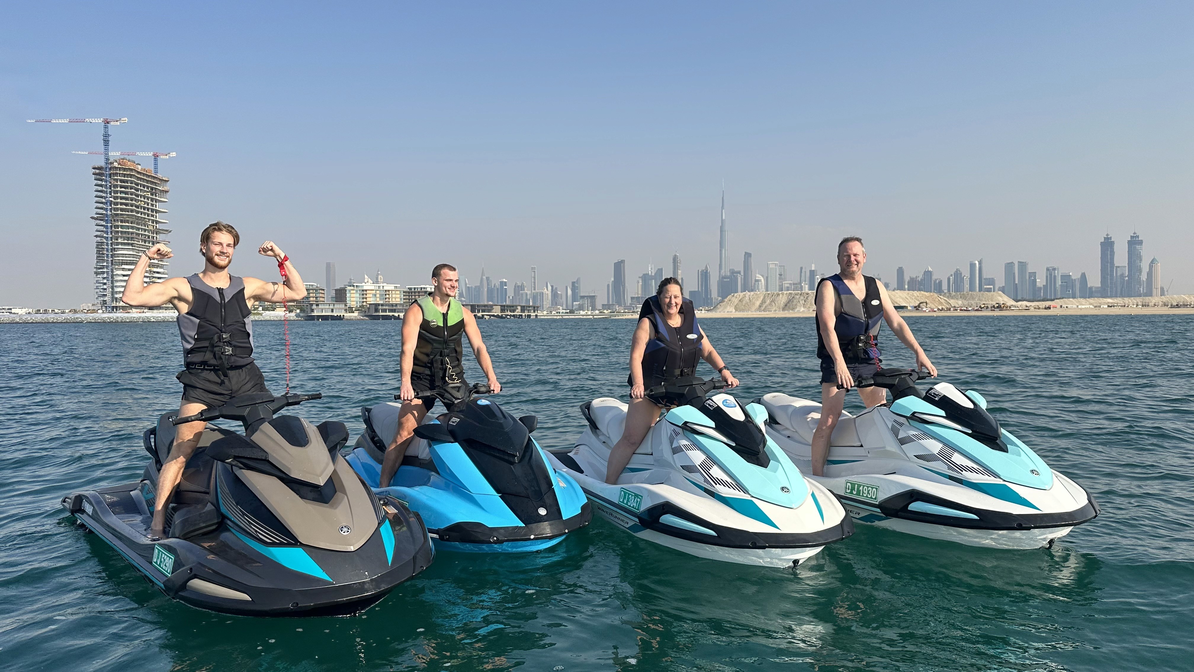 A group of four people riding jet skis on the water, with the Dubai skyline visible in the background.