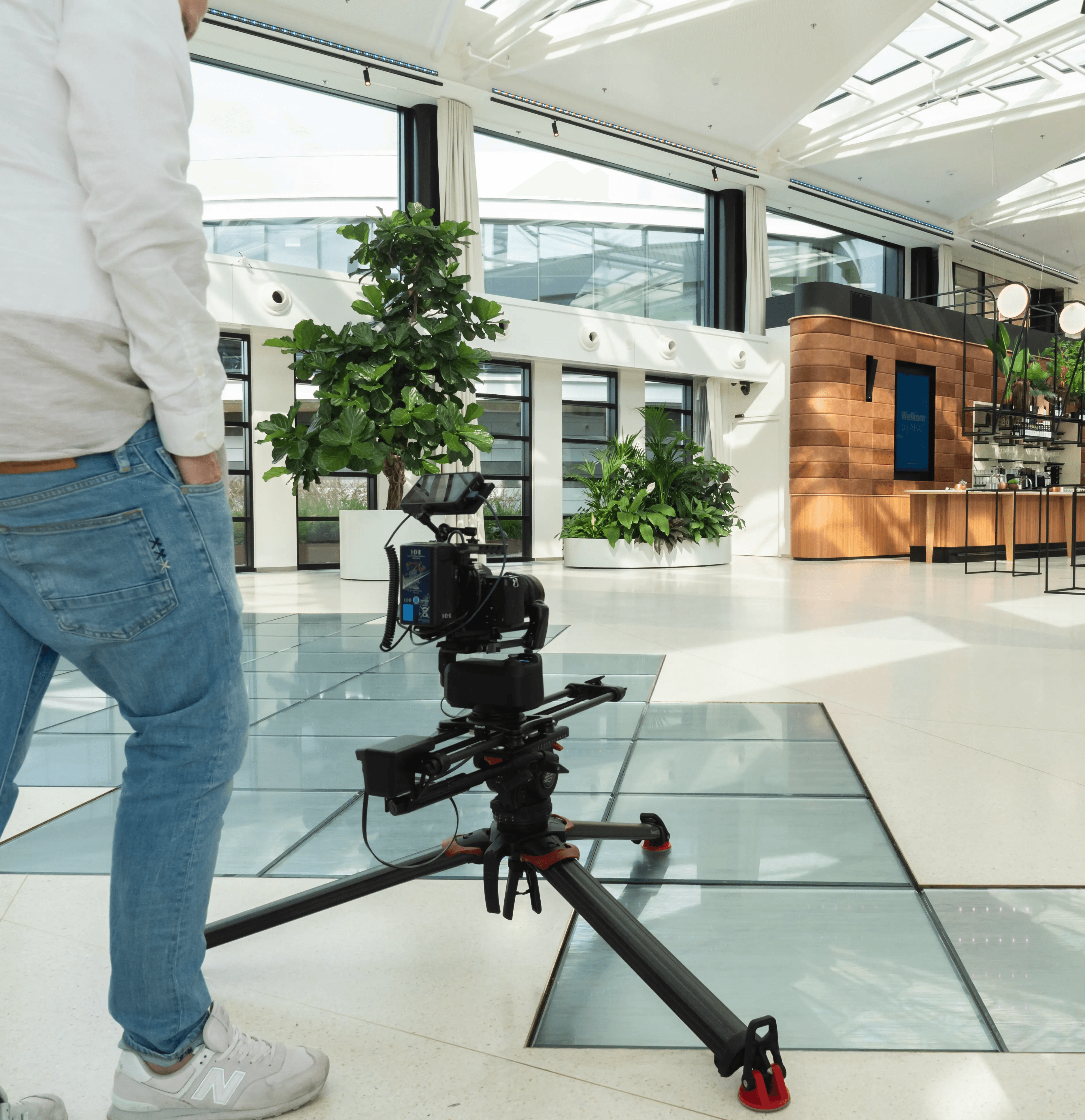 Een persoon stelt een professionele camera af op een tripod dolly in een ruime, moderne lobby met grote ramen, weelderige binnenplanten en strakke houten accenten, wat de heldere en eigentijdse architectonische vormgeving benadrukt.