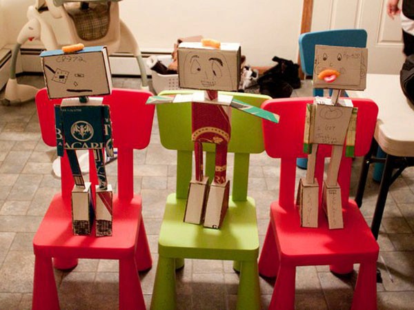 Three cardboard robots stand on brightly colored children's chairs: a red chair on the left, a green chair in the center, and a red chair on the right. Each robot is constructed from cardboard boxes and features simple facial expressions and shapes. The chairs are placed on a tiled floor, with other chairs and a table visible in the background.