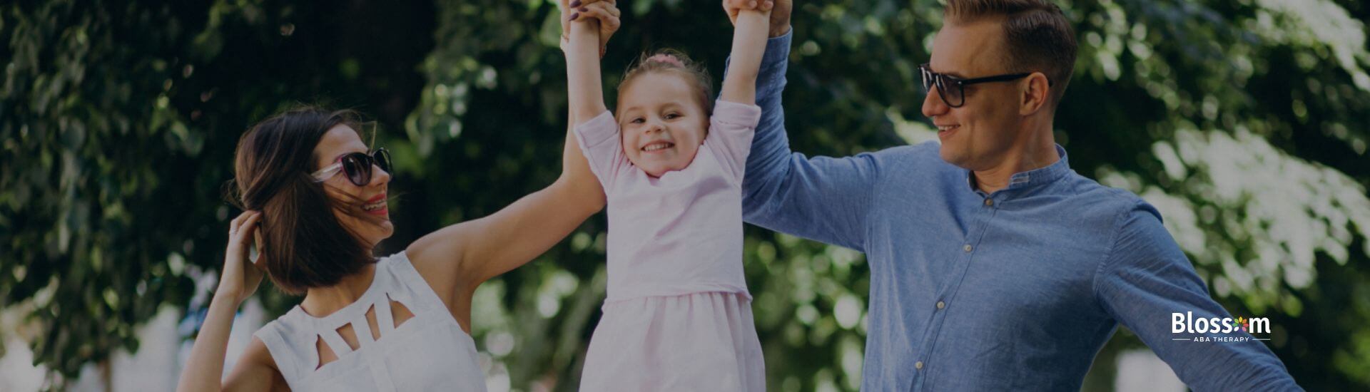 Parents lifting their smiling daughter with autism by the arms while walking outside together.