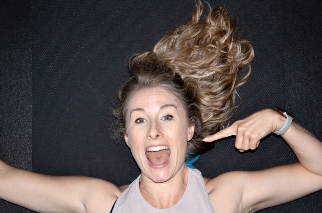 Heather Hausman lying on a dark textured surface, pointing at her smiling face, wearing a sleeveless top and a fitness tracker.