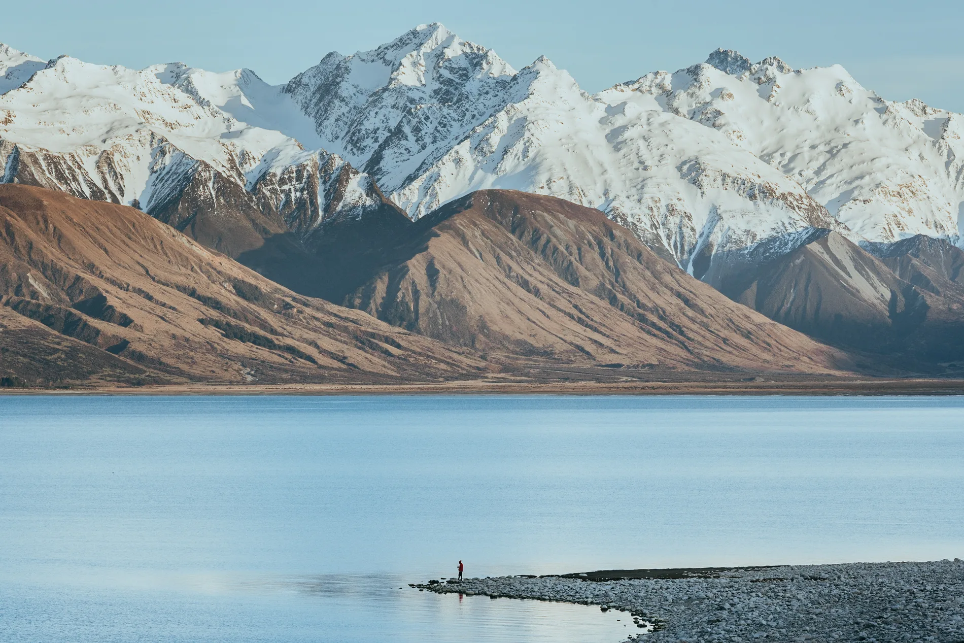 Snow-capped mountains above a pale blue lake, a lone figure on the shingle spit