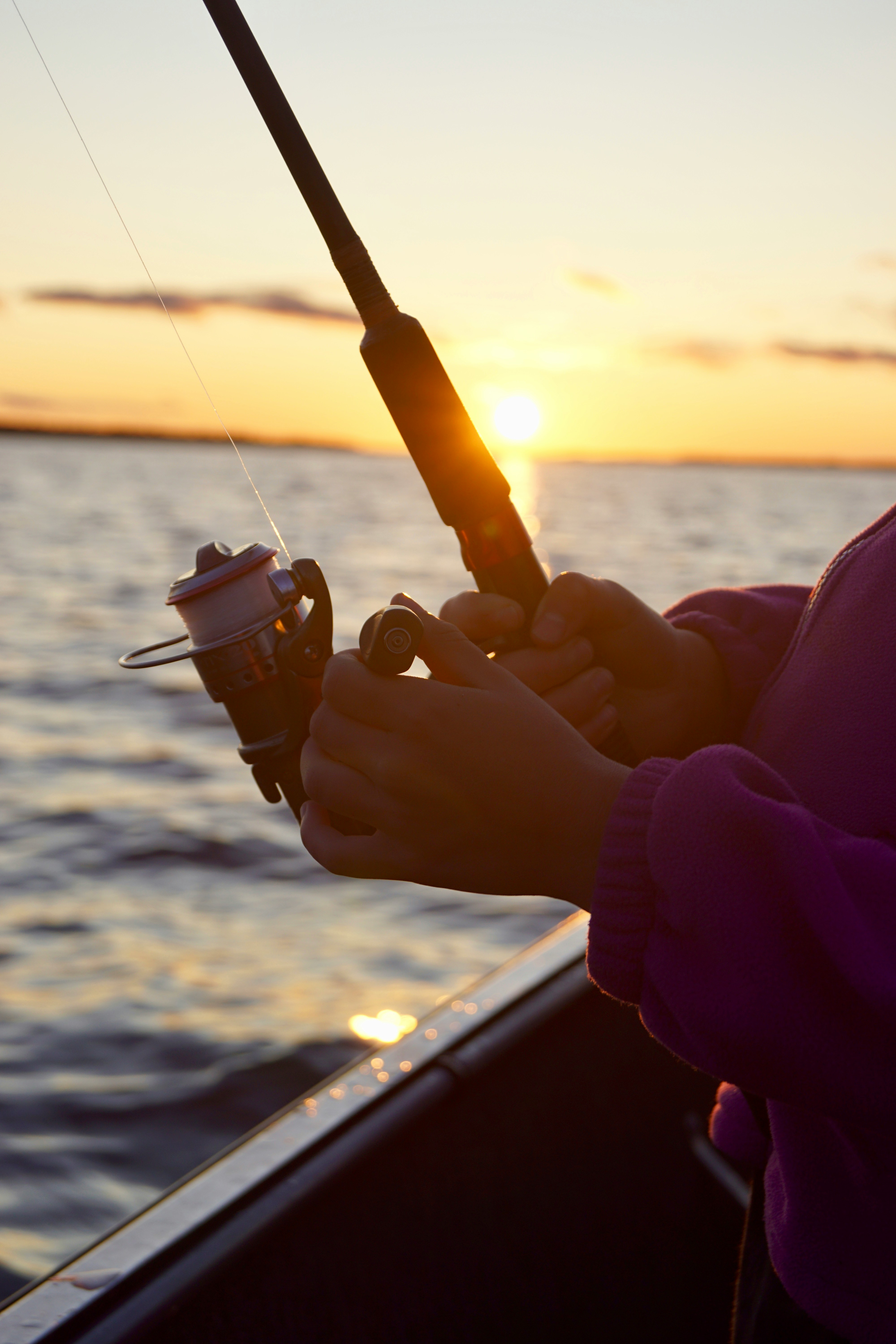 A person in a purple jacket holds a fishing rod against a backdrop of a tranquil sunset over a vast, rippling lake, with the sun casting a golden glow across the water.