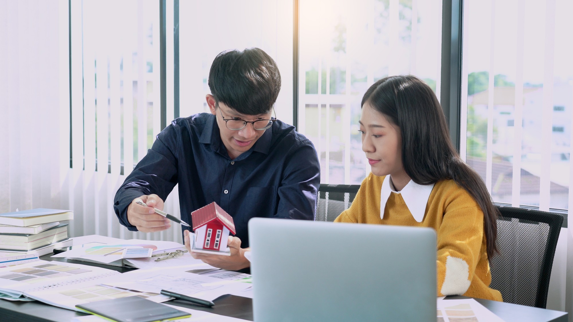 Real estate agents reviewing investment data on a laptop while discussing a model house, symbolizing property sales strategy.