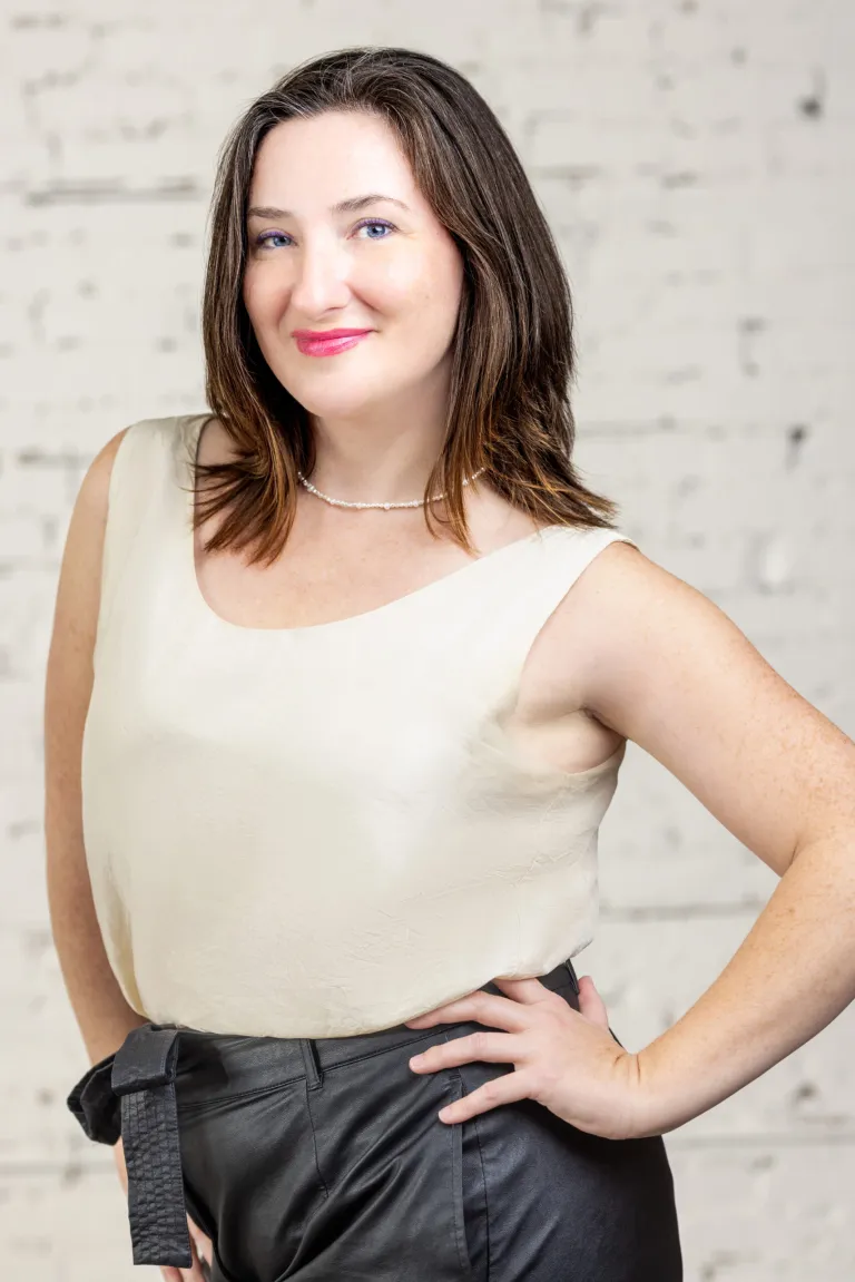 Image of Woman smiling with hand on hip standing in front of white brick wall background confidently.