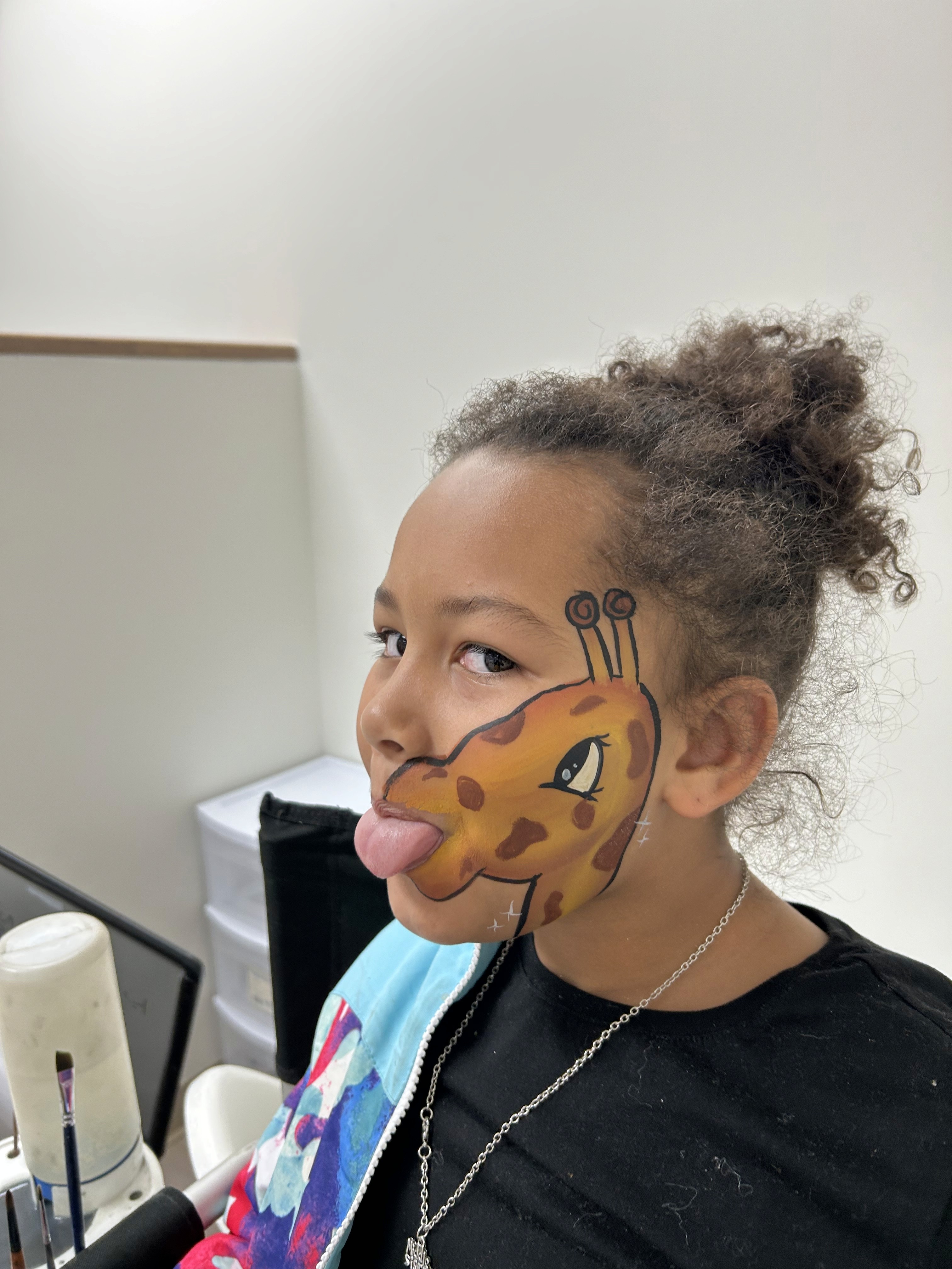 A young girl with her face painted as a cheerful giraffe sticks out her tongue playfully. The detailed face painting highlights vibrant brown spots and long giraffe features, creating a fun and whimsical look.