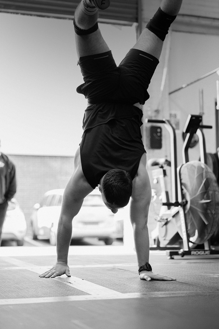A muscular man smiling while performing a hanging leg raise on gymnastic rings in a bright fitness studio.