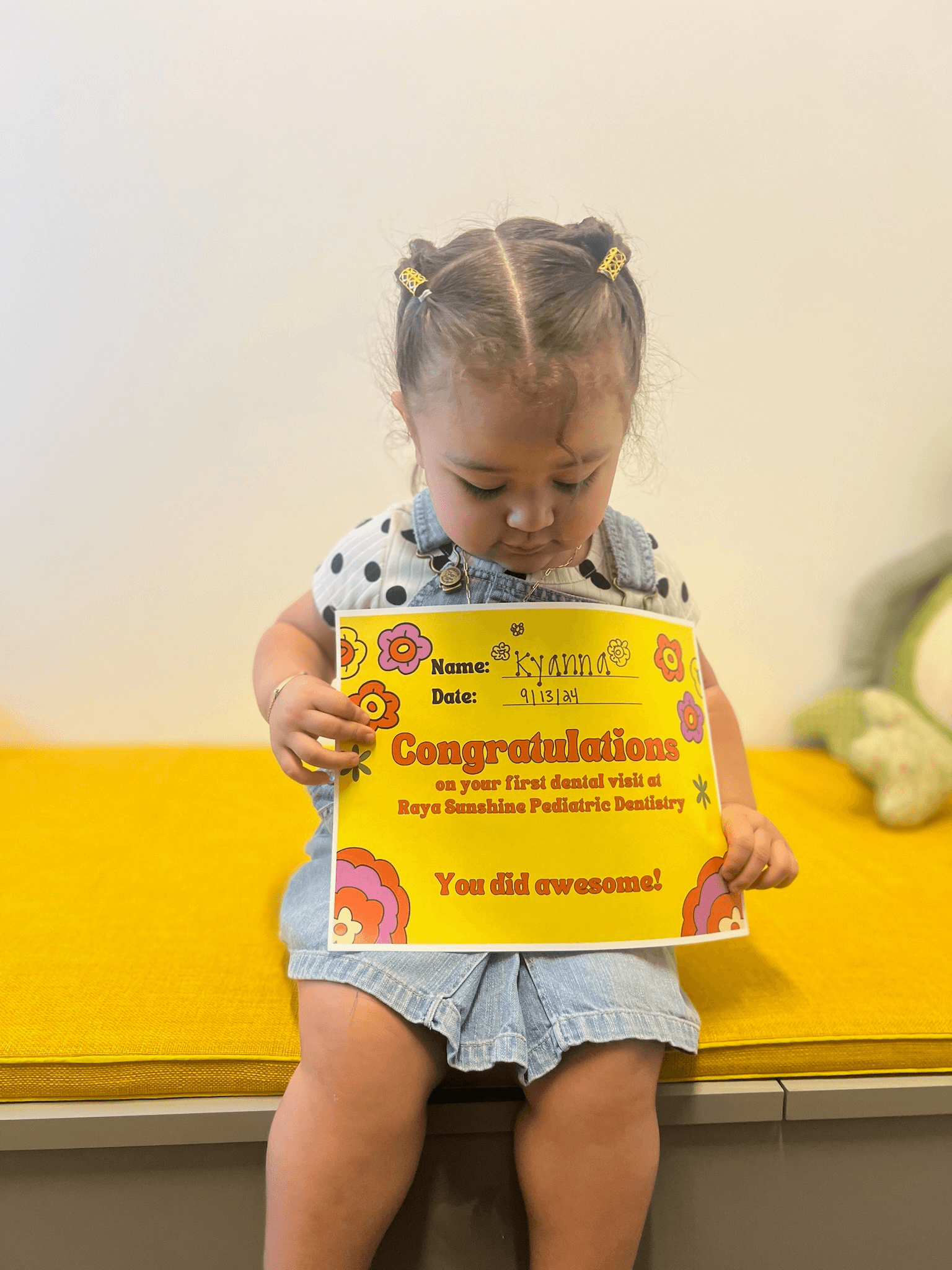 A toddler sitting on a bench holding a Raya Sunshine Pediatric Dentistry sign, looking down at it