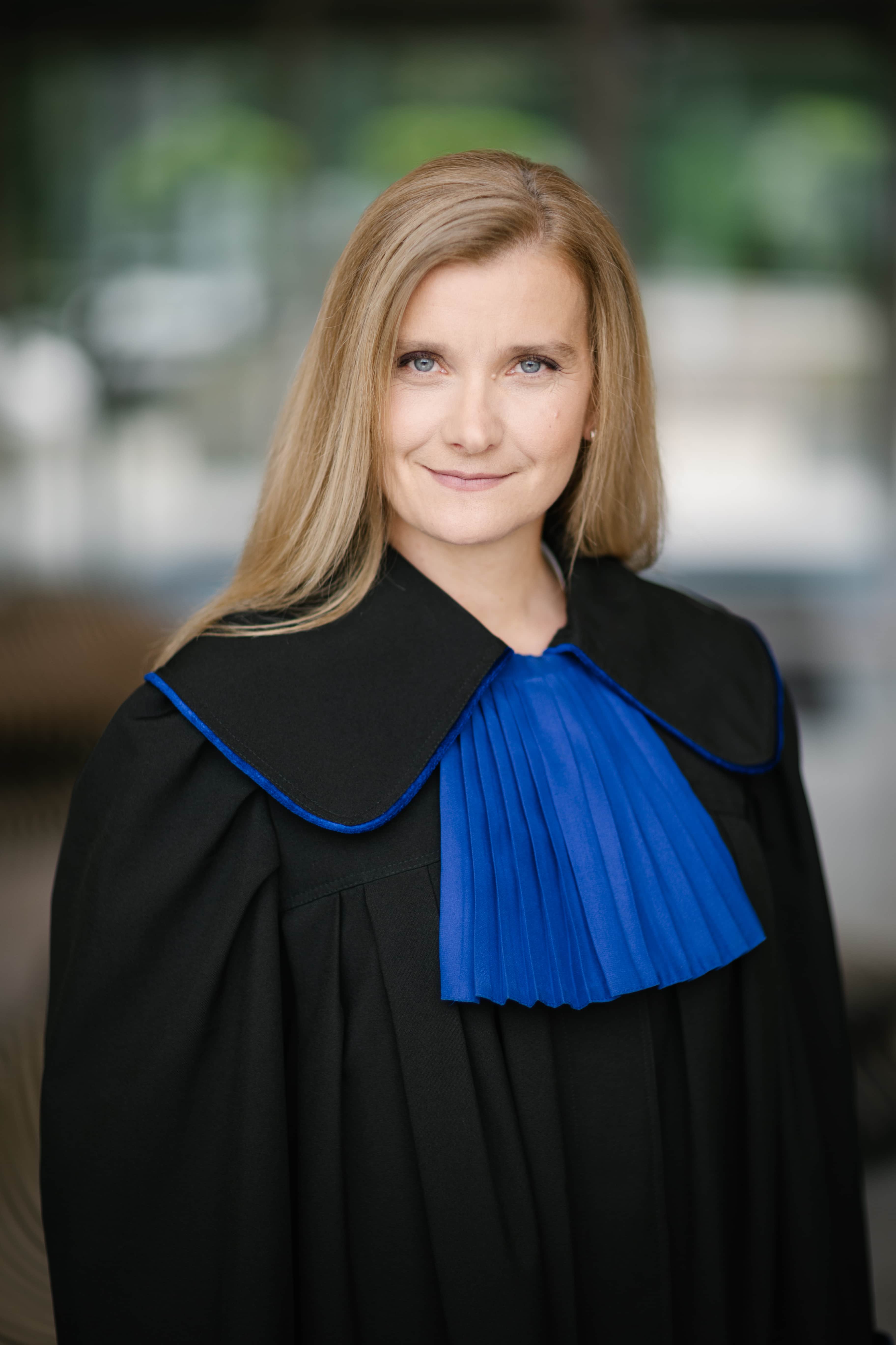 Kobieta o długich blond włosach ubrana w czarną togę akademicką lub sędziowską z niebieskim plisowanym kołnierzem stoi w pomieszczeniu, patrząc pewnie w kamerę. Tło jest delikatnie rozmyte.