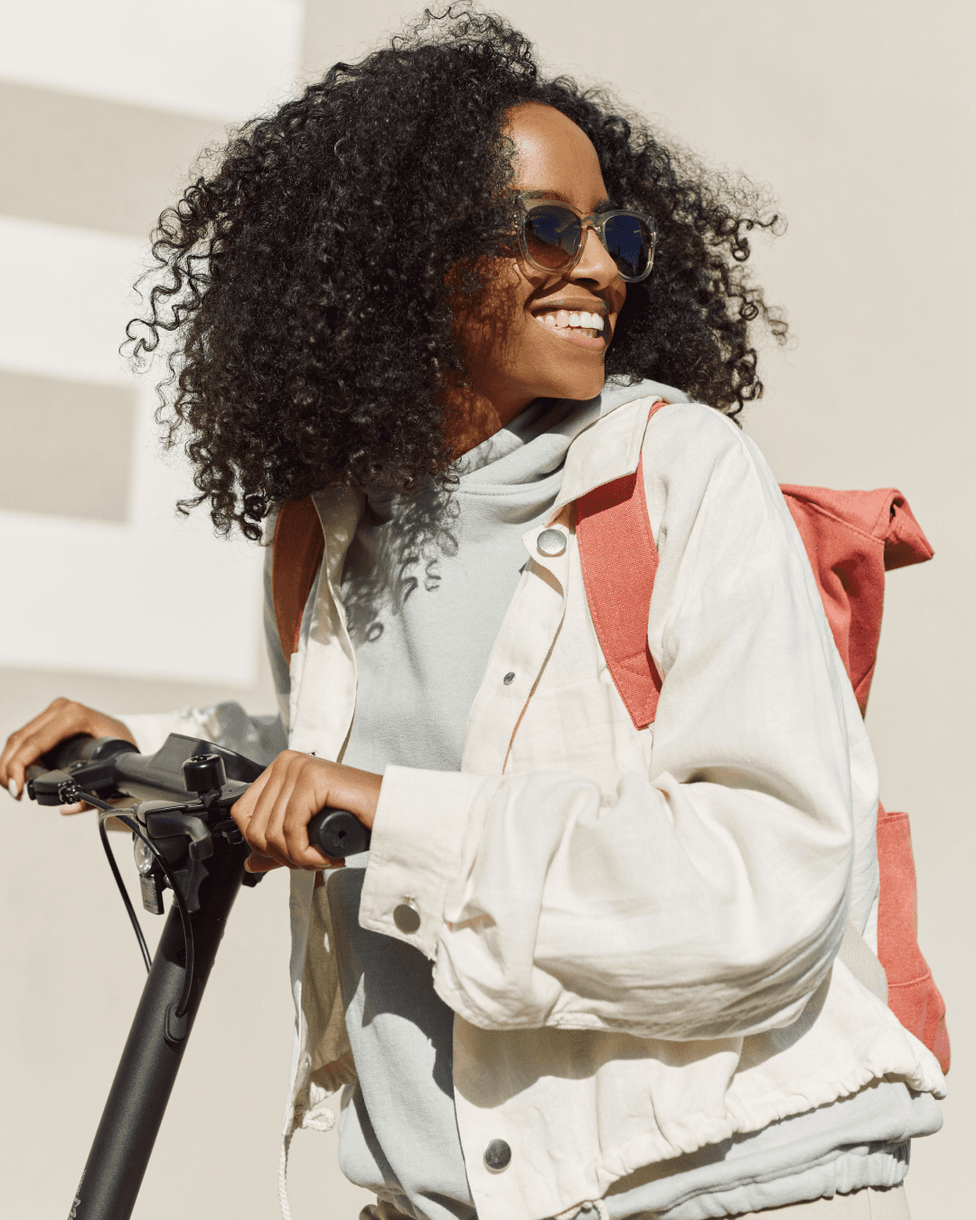 Woman on scooter smiling and wearing smart sunglasses. White jacket, orange backpack.