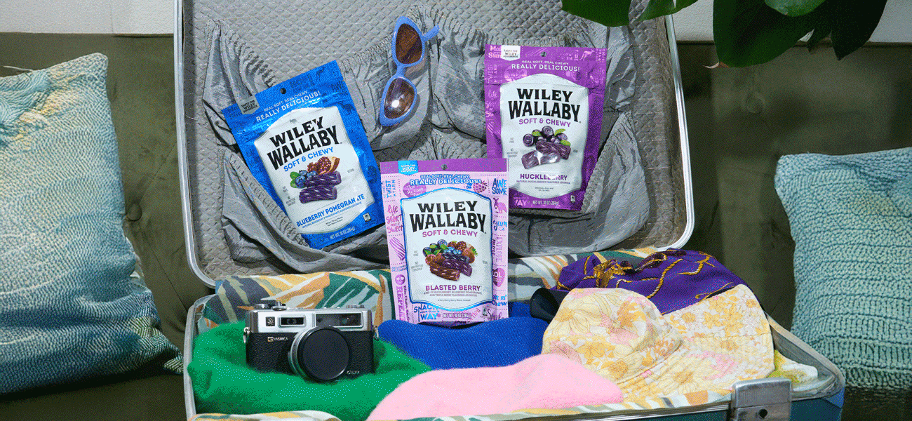 Wiley Wallaby soft and chewy candy bags arranged inside an open suitcase with clothing, camera, and accessories, styled as a travel-themed lifestyle scene.
