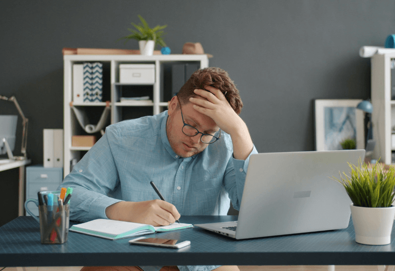 Man sitting at a desk looking mentally fatigued while writing in a notebook in a home office setting.