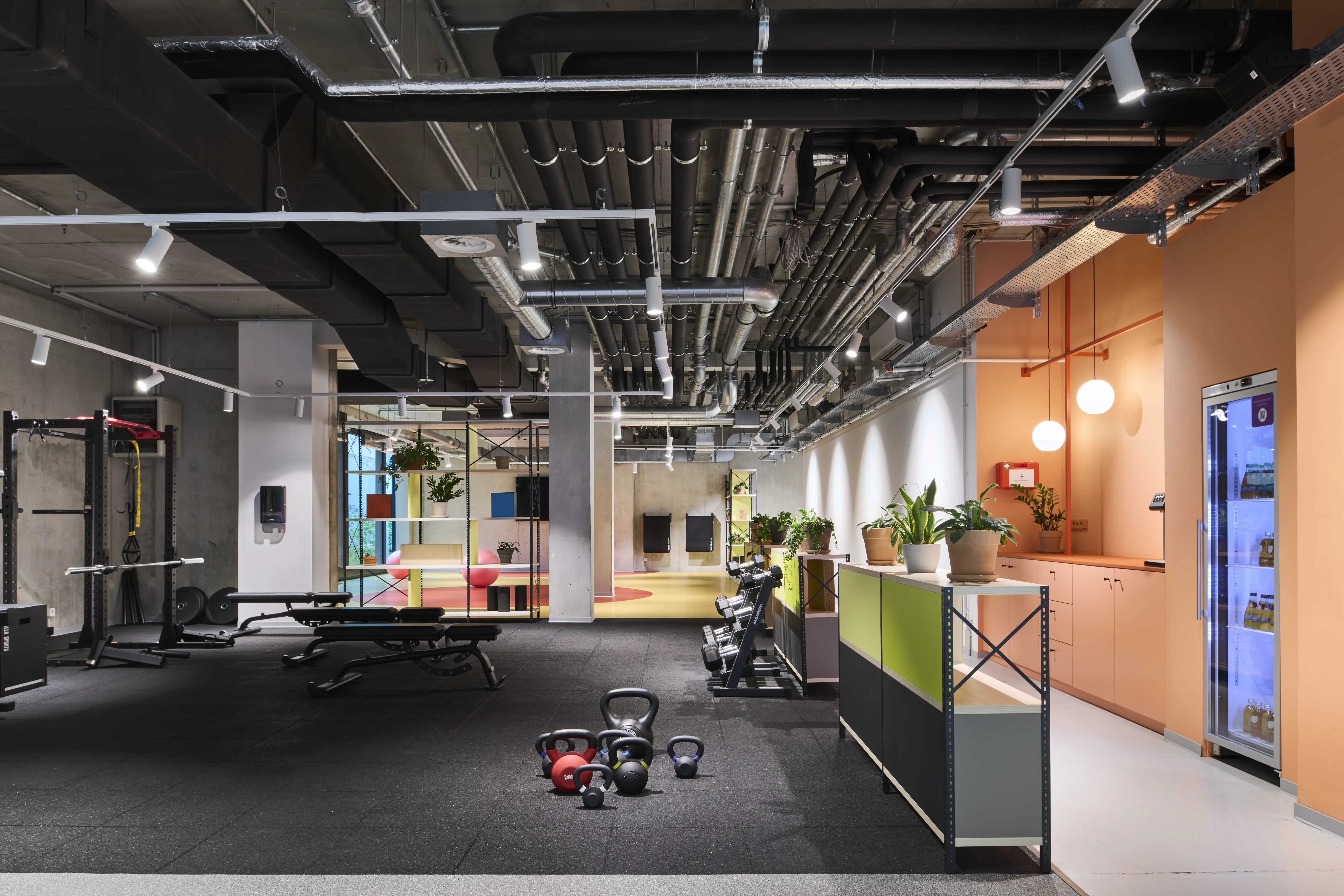 Overview of the HELLOFRESH GYM with black rubber training floor, kettlebells, a green-grey equipment sideboard, apricot kitchen wall and exposed ceiling pipes.
