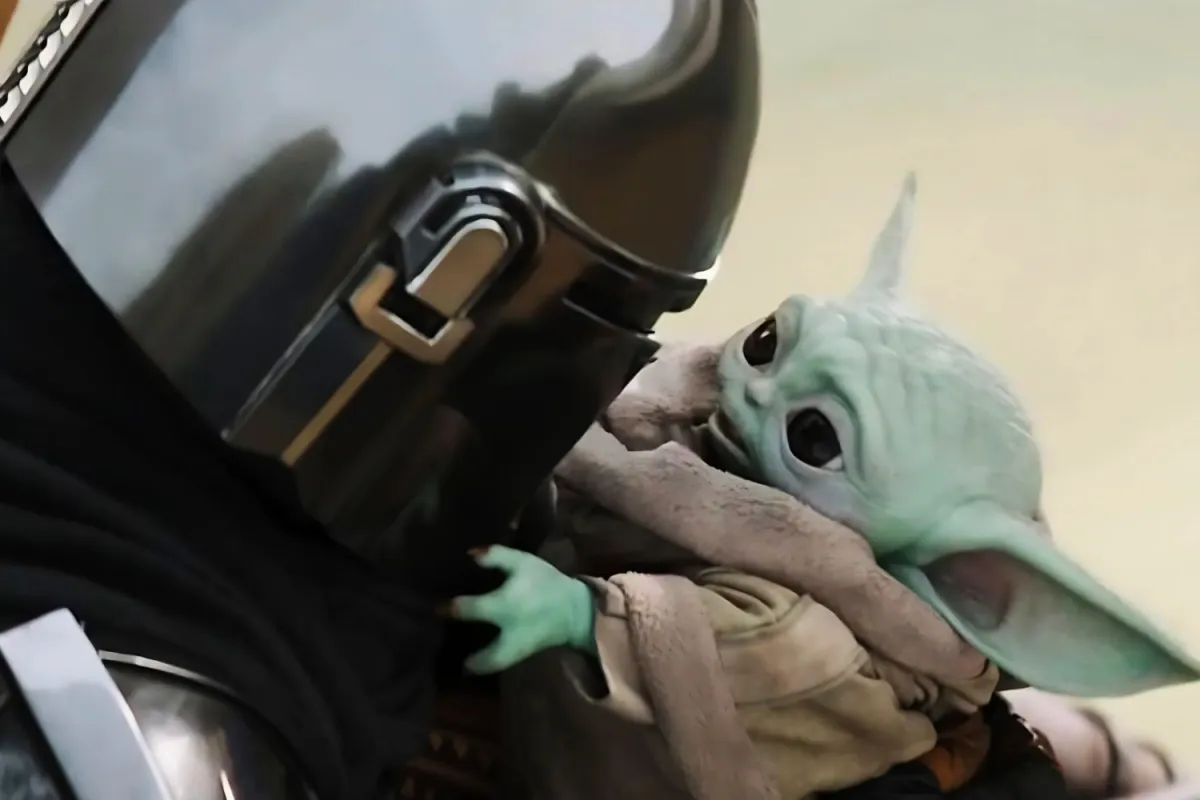 Grogu clings to Din Djarin’s armored chest, gazing up at him with joy and relief. Din holds him close, their reunion marking the restoration of their unbreakable bond after Grogu’s brief time away with Luke Skywalker.