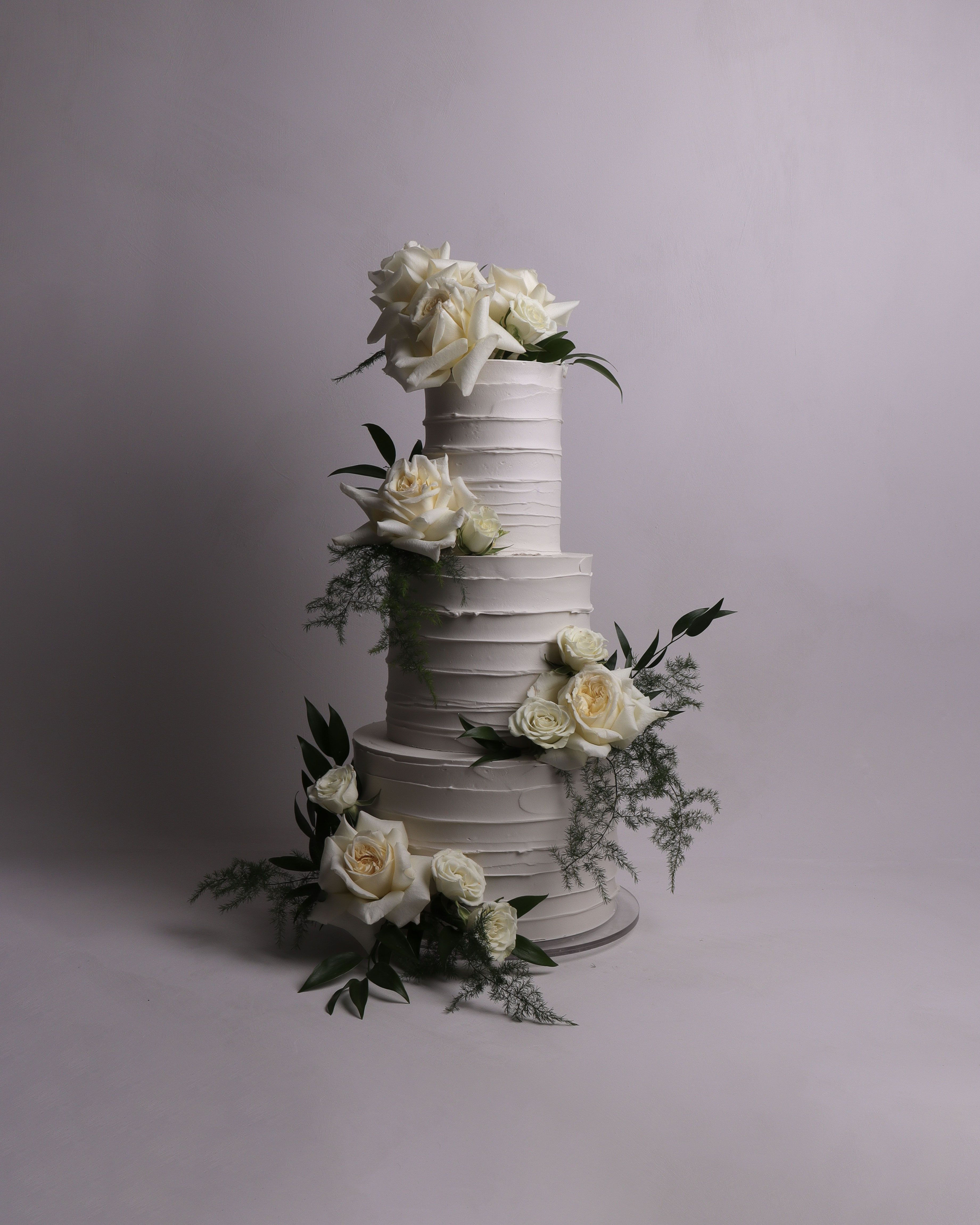 Three tier wedding cake with textured buttercream finish styled with fresh white roses and soft greenery arranged vertically across the tiers
