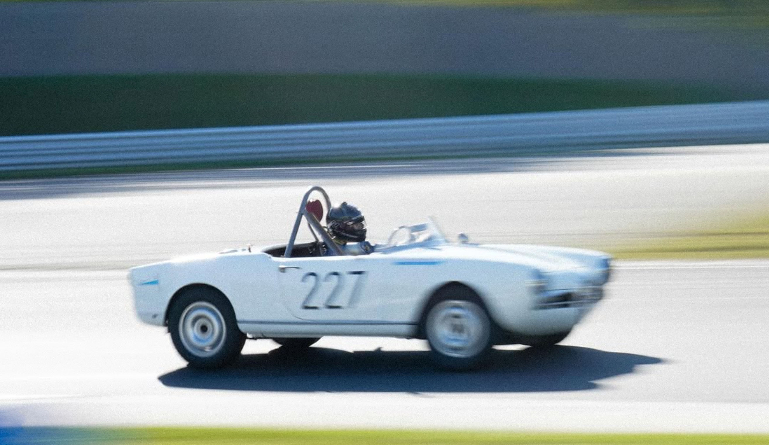 A white classic convertible car speeds around a racetrack with a driver inside, capturing a dynamic motion scene.