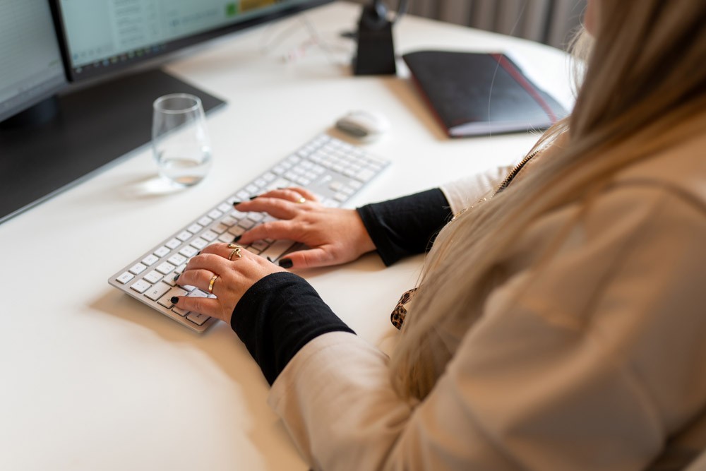 Person arbeitet an einem PC, neben der Tastatur liegt ein Notizblock auf dem Schreibtisch.