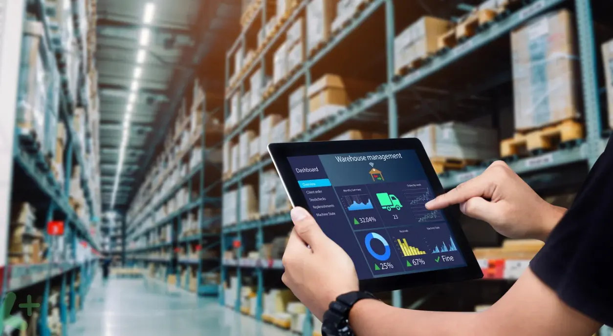 An operator consulting a tablet with a warehouse management dashboard in an aisle of shelving.