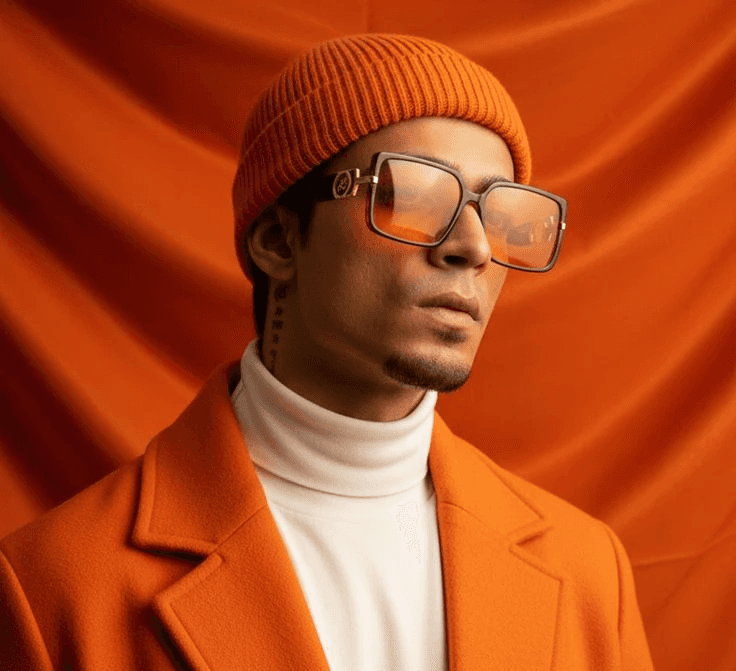 A person in an orange beanie, large sunglasses, and a coat poses against an orange backdrop. They wear a white turtleneck, exuding a stylish, confident vibe.