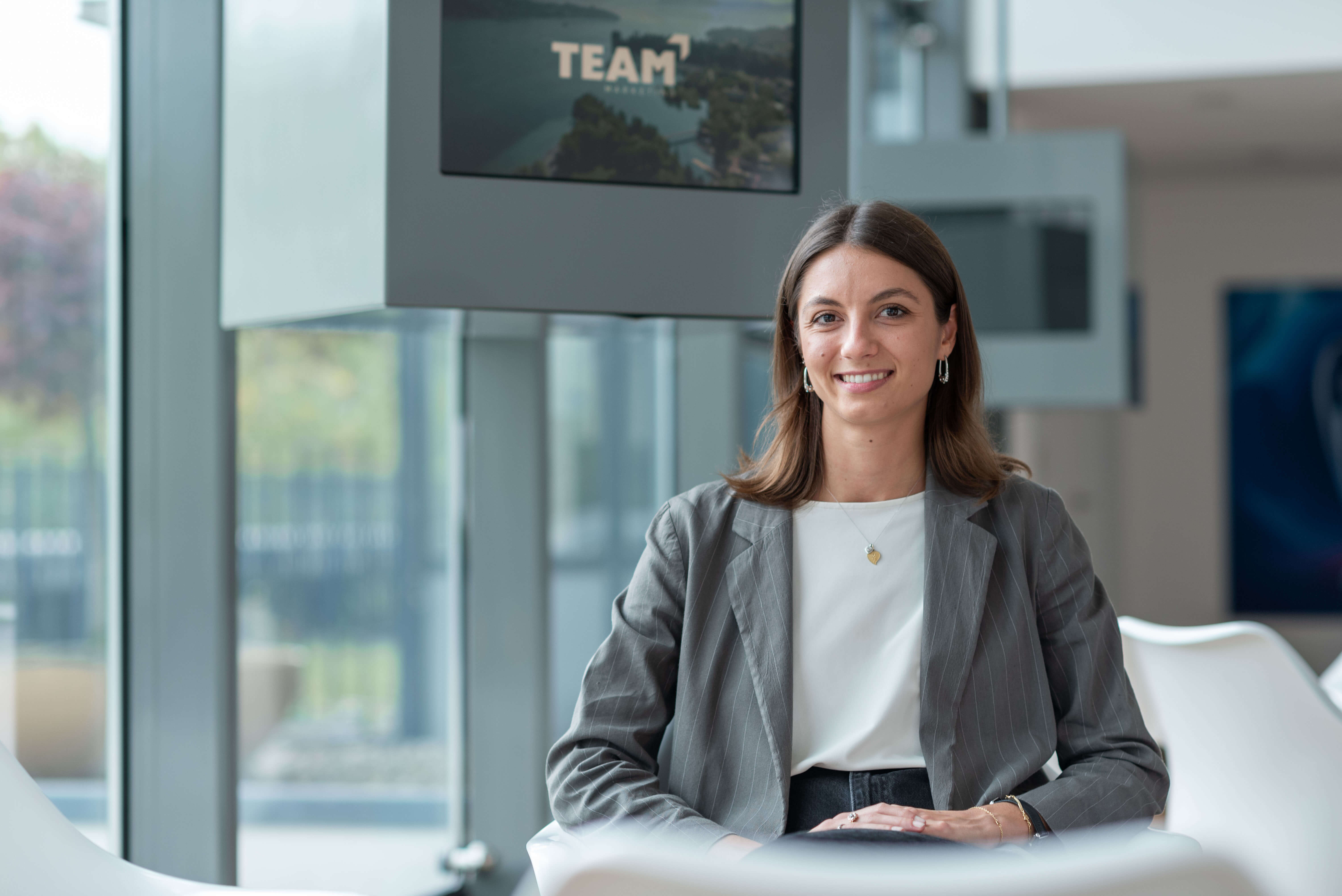 Female Team member seated confidently in a modern office space, with a Team logo visible in the background.