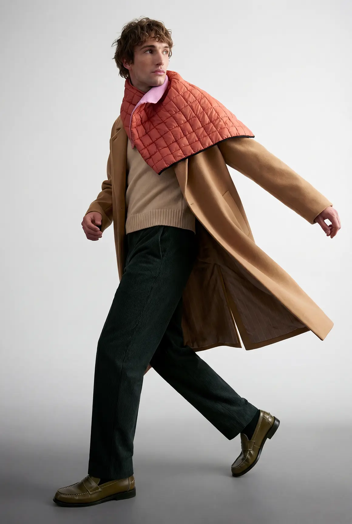 A person confidently walking is dressed in a stylish camel overcoat paired with a quilted orange scarf, dark trousers, and olive-green shoes, showcasing high-end visuals suitable for an AI production project in a fashion brand campaign.