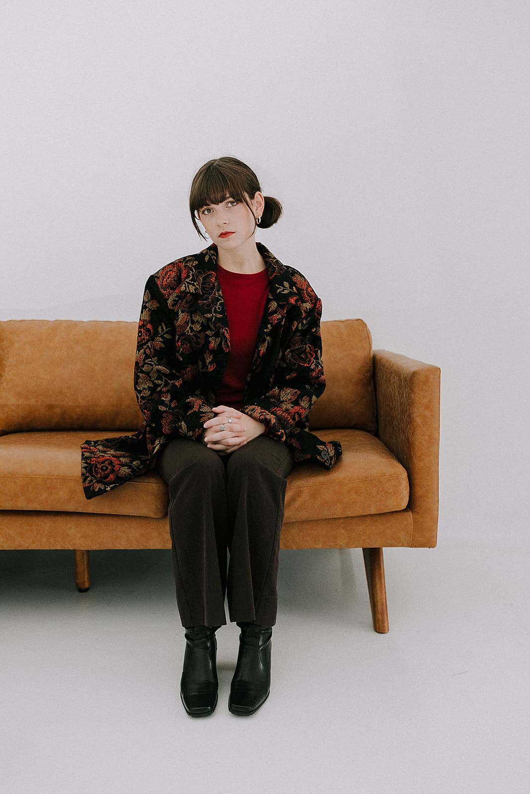 A model sitting on a tan couch, dressed in a floral jacket and brown pants, striking a serious pose during the 90s vintage photoshoot at Revelator Studio, Shreveport.