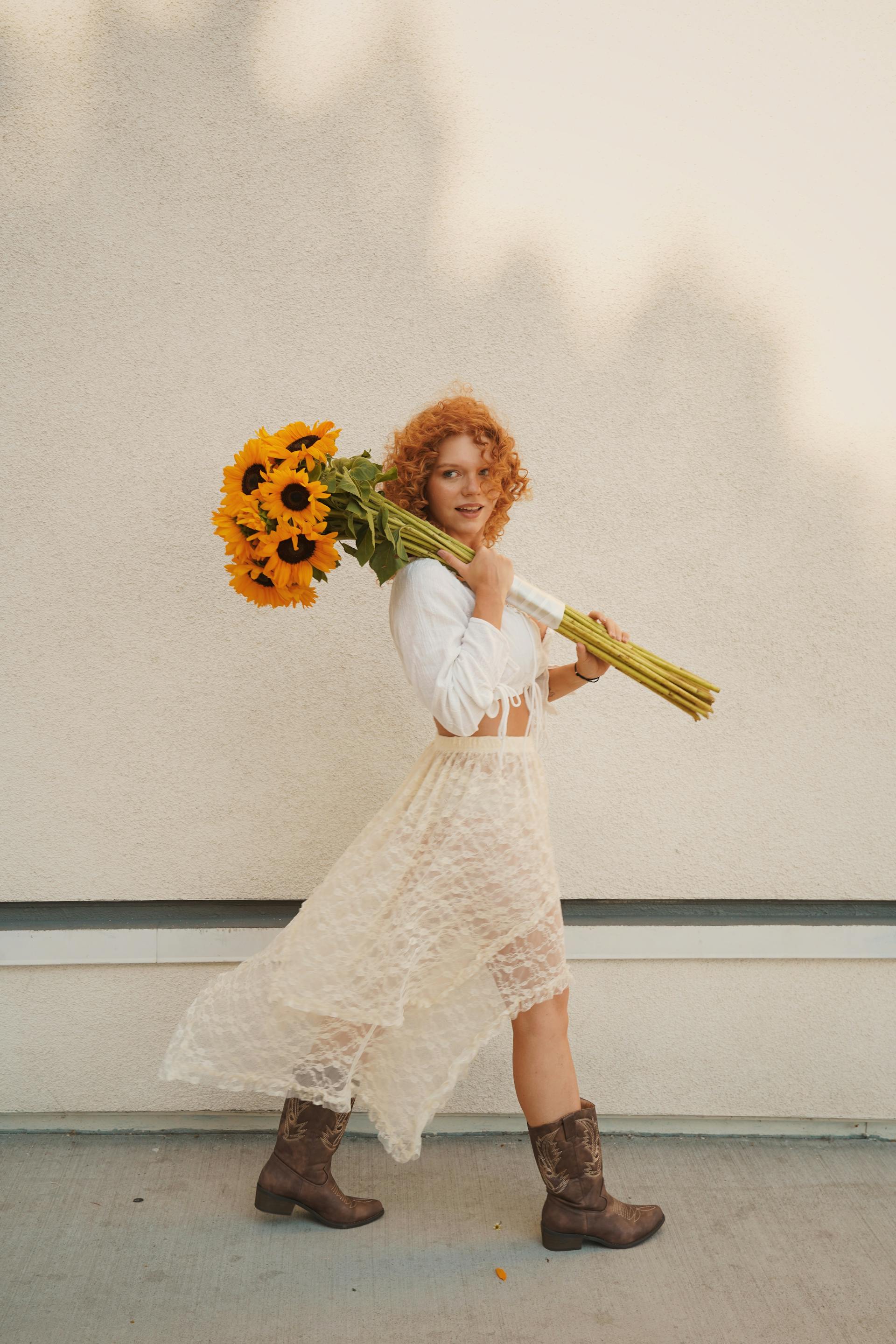 A woman with curly red hair, wearing a white top, lace skirt, and brown boots, walks while carrying a large bouquet of sunflowers over her shoulder.