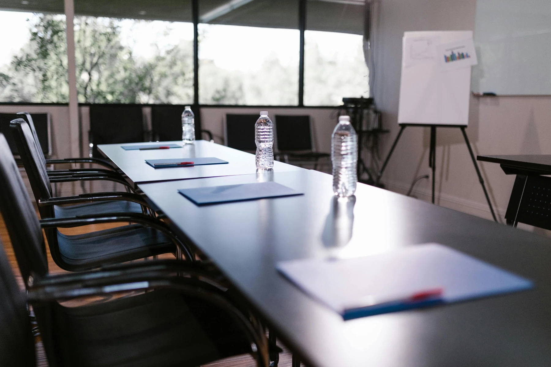 A meeting room setting with a long table, water bottles, blue folders, and pens. Large windows provide natural light.