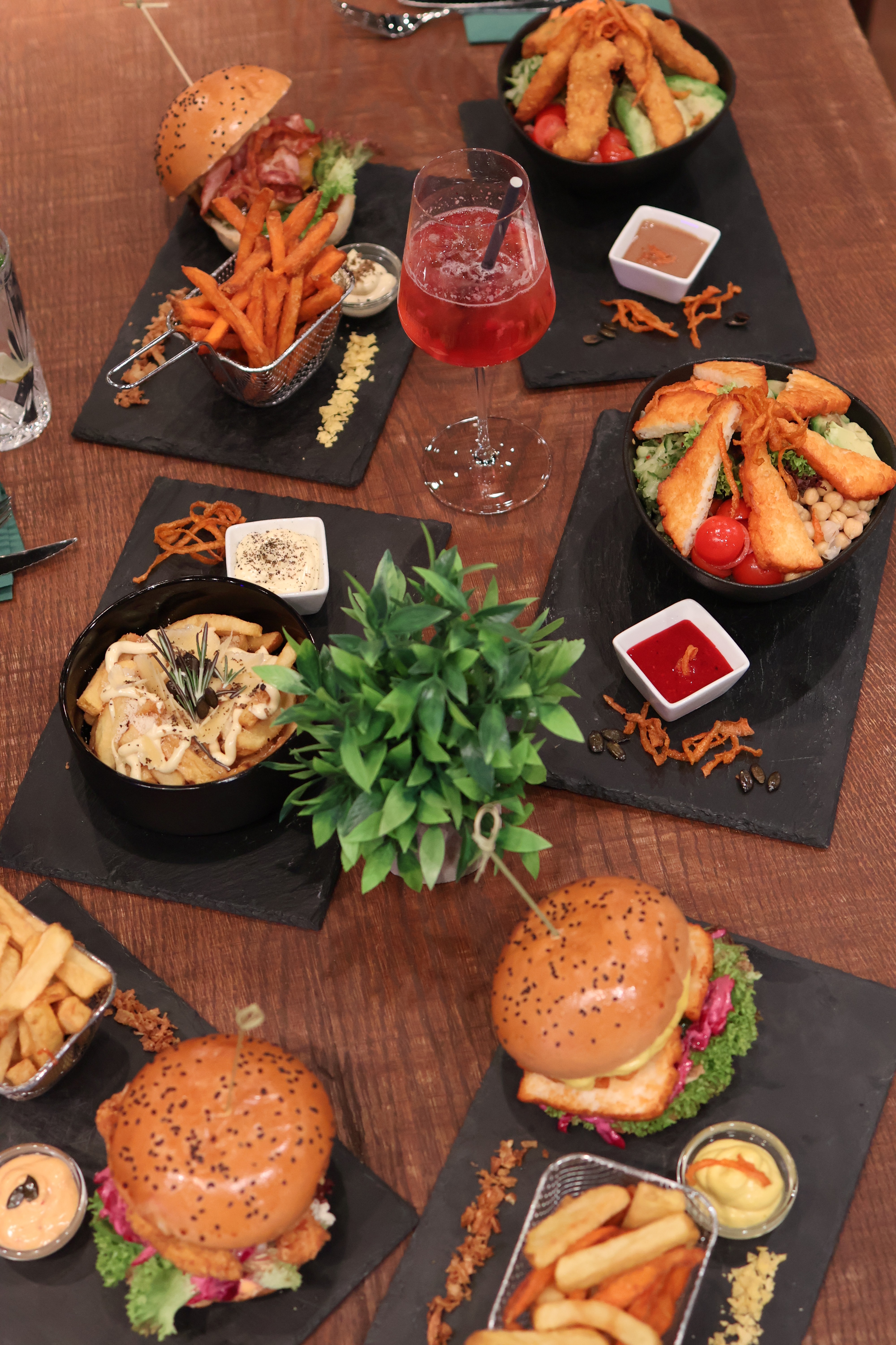 Overhead view of a kAlps Foodbar table spread with burgers, crispy fries, fondue dishes, a red cocktail, dipping sauces, and fresh garnishes arranged on slate serving boards.