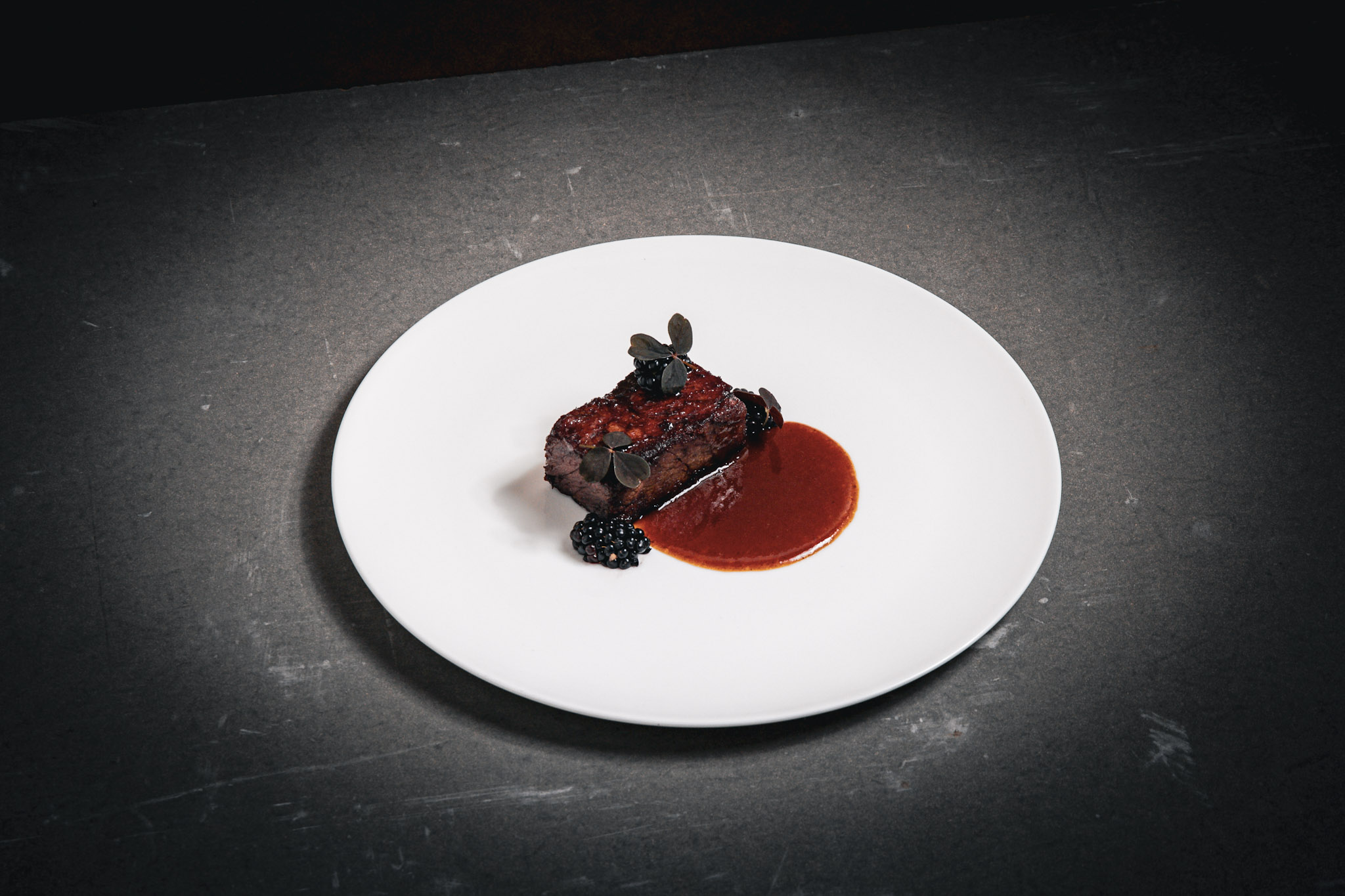 Beef short rib with reduced sauce and whole blackberries, plated simply with minimal garnish and dark, glossy finish.