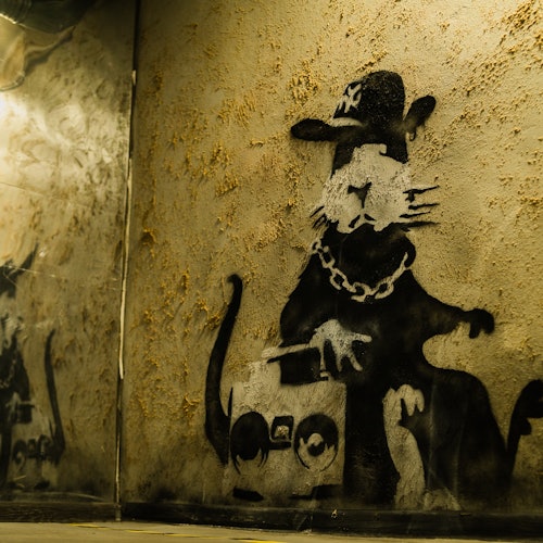 Un graffiti de un gato usando un sombrero y una cadena, sosteniendo un boombox, está pintado en una pared beige con textura.