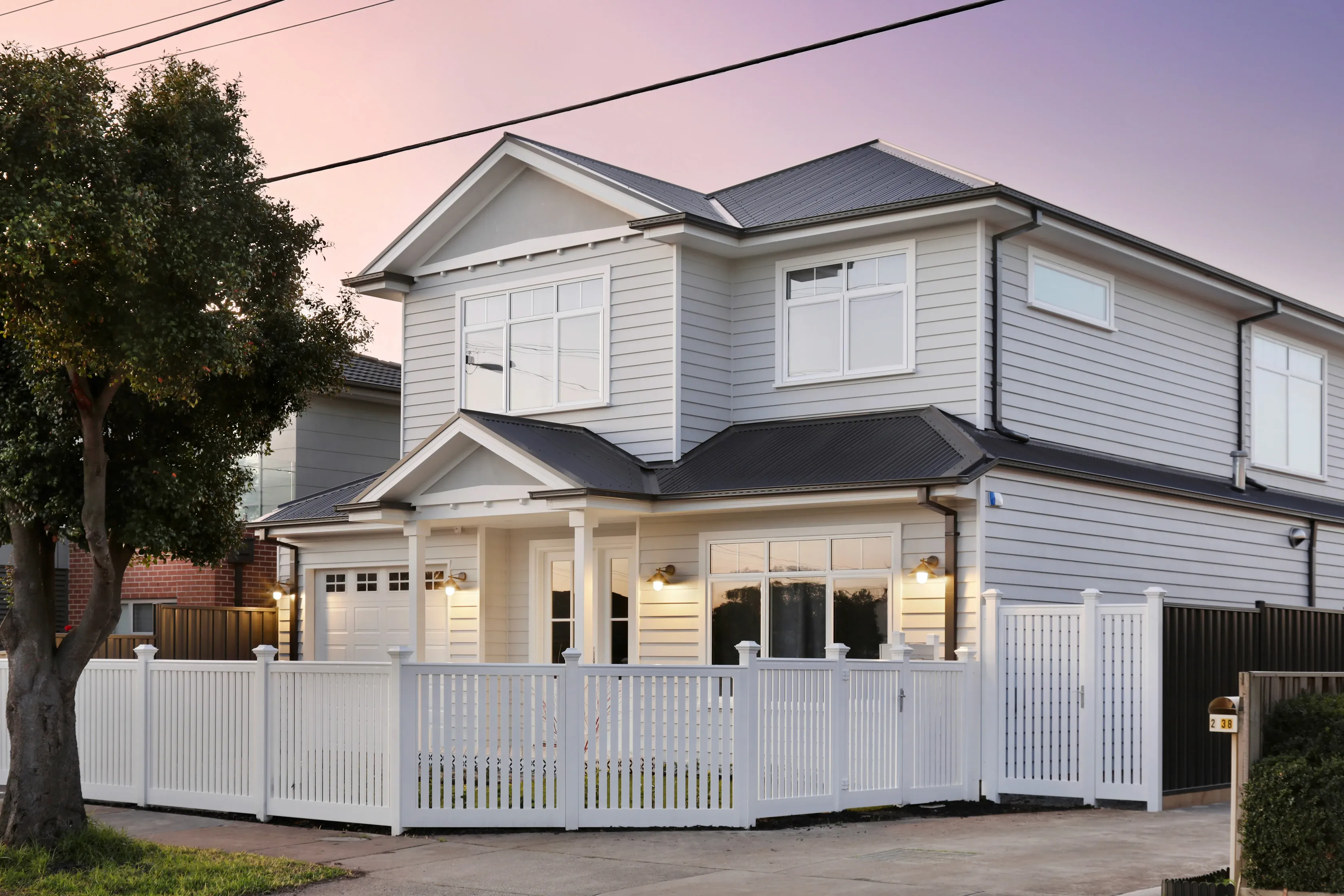 Front of the Lalor Residence at dusk, a two-storey pale weatherboard Hamptons home with a gabled entry portico, dark tiled roof, carriage lights, a white picket fence and gate beside a tree