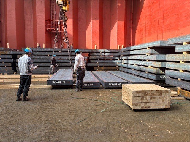 Stevedores loading steel slabs inside a vessel’s cargo hold using a crane during port operations.