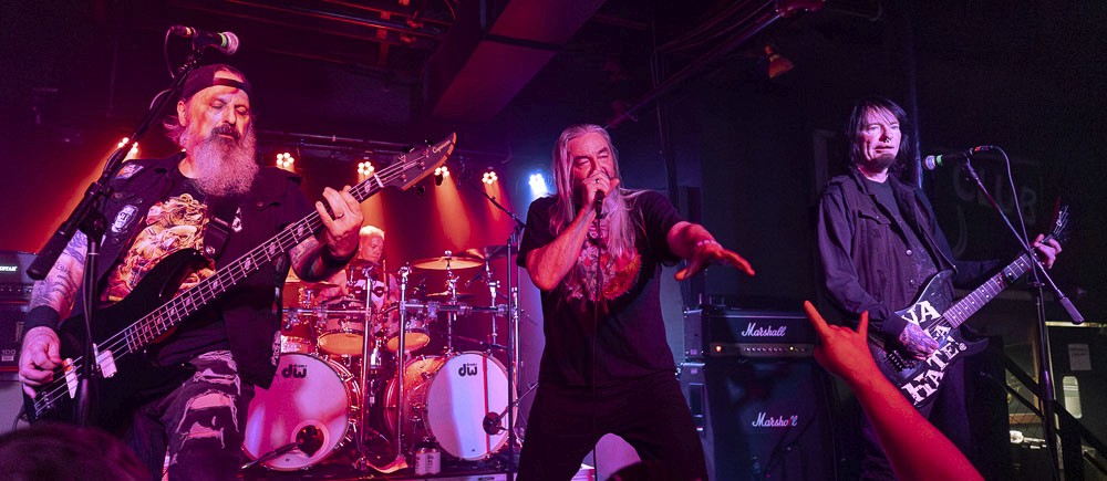 A rock band performing a high-energy live show on stage, featuring a singer at the microphone and a drummer behind a full kit under intense pink and red stage lights.