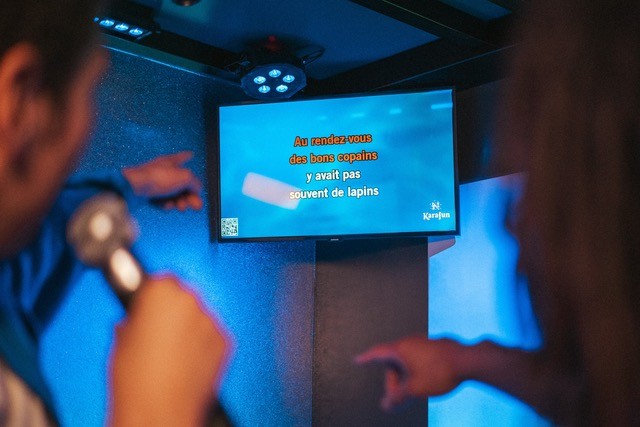 A young person with curly hair smiles at the camera while sitting in front of a laptop against a dark background.