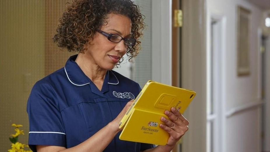 Nurse in uniform using a yellow tablet while standing in a hallway inside a care facility.