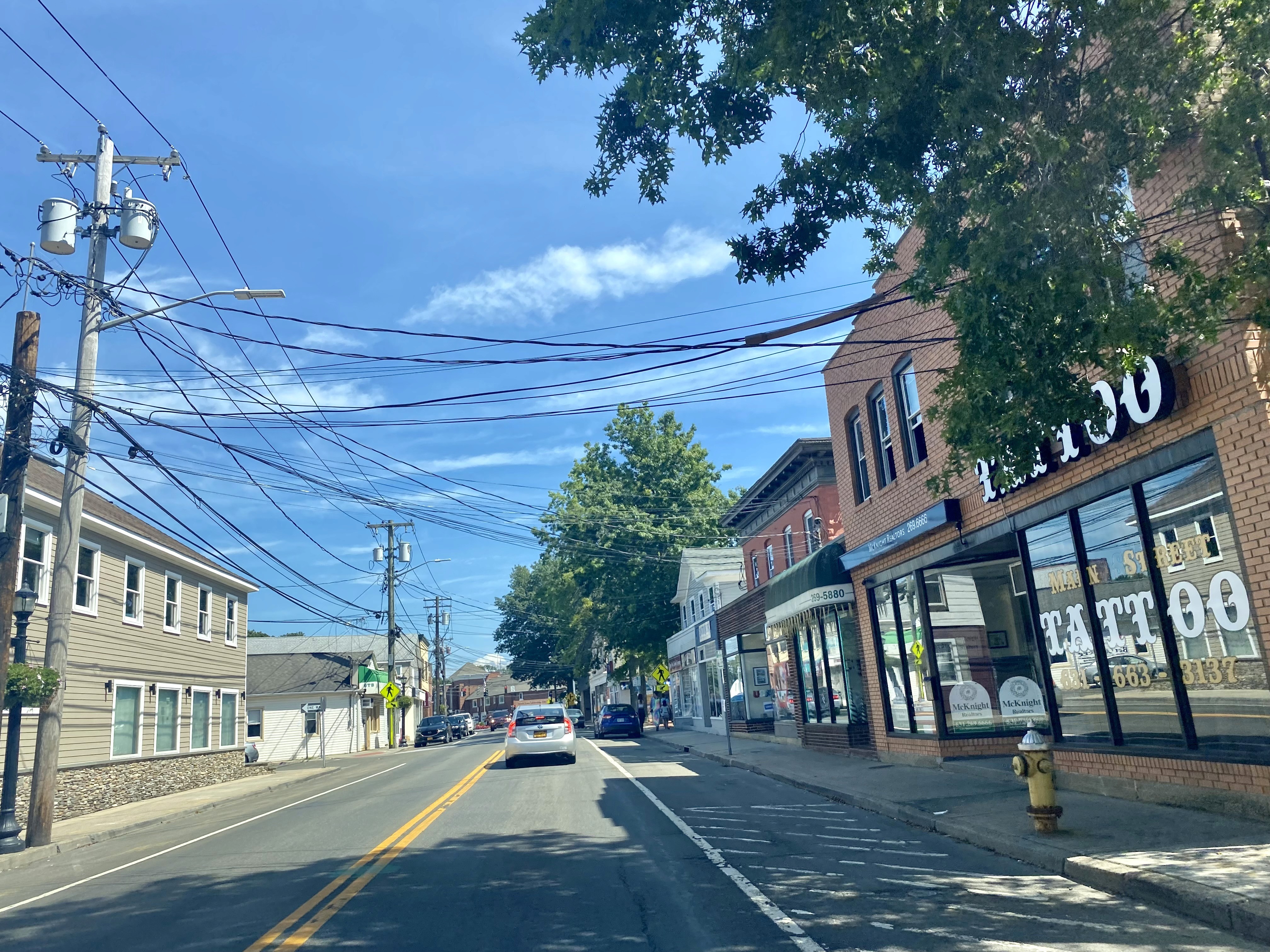 A sunny street scene with blue skies, trees lining the road, and buildings on the side.