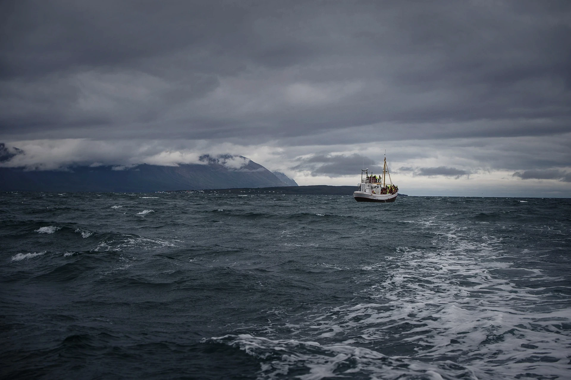 Small fishing boat on rough sea with dark storm clouds and distant coastline.
