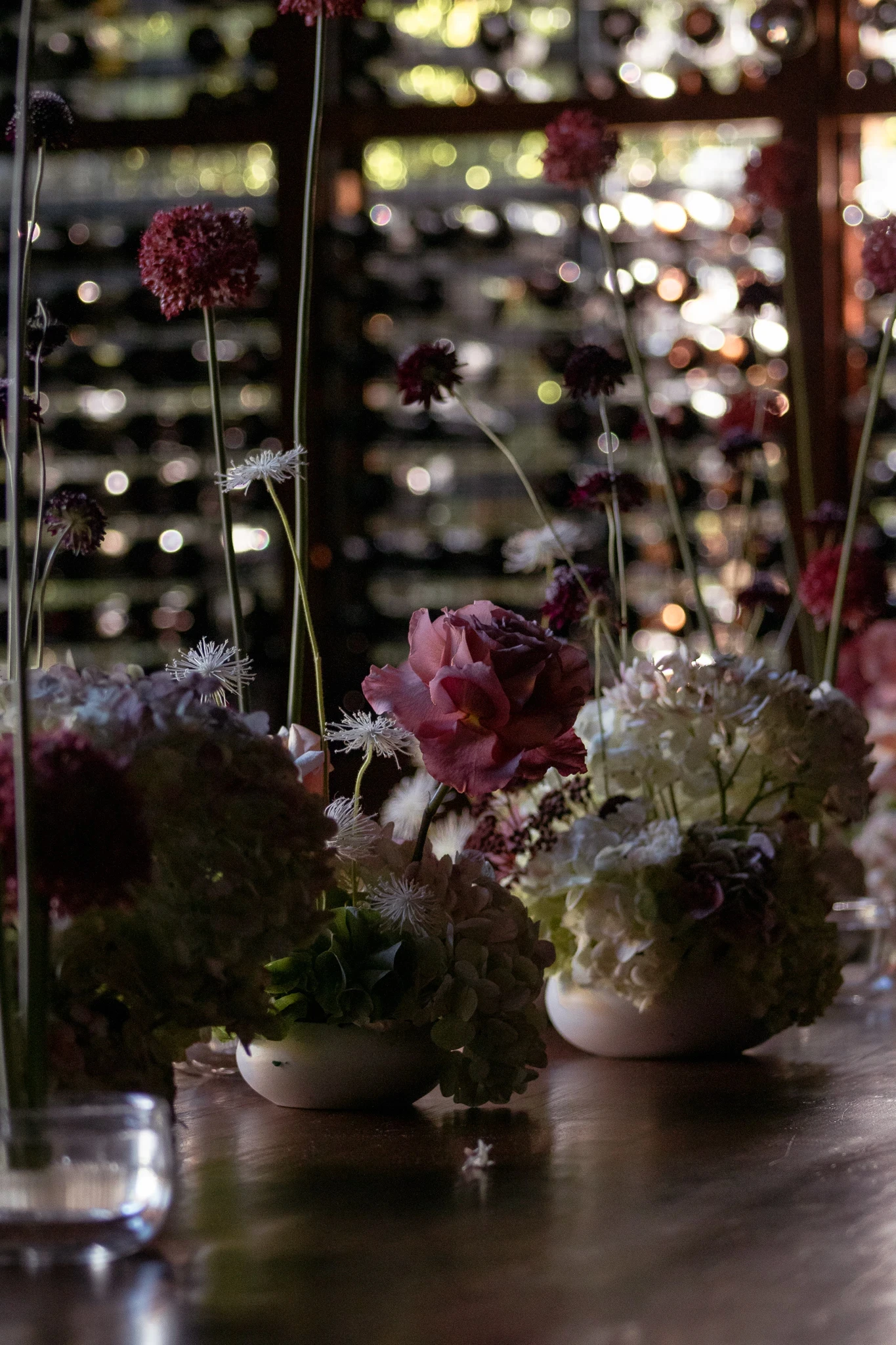 Centros de mesa con rosas malva y hortensias blancas frente a una imponente cava de vinos de madera oscura.