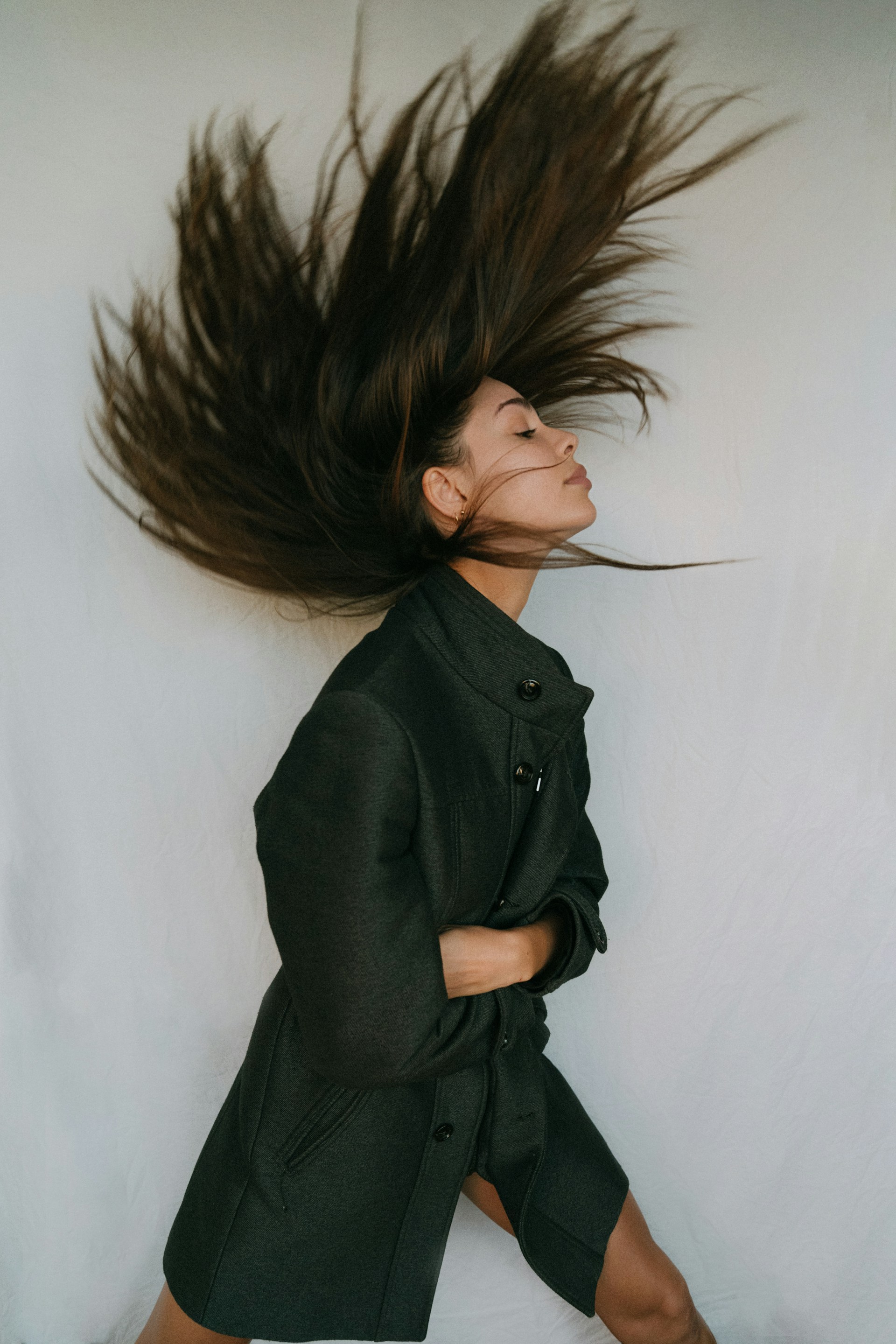 A woman in a dark coat with her long hair fanned out upwards in motion against a plain white backdrop.