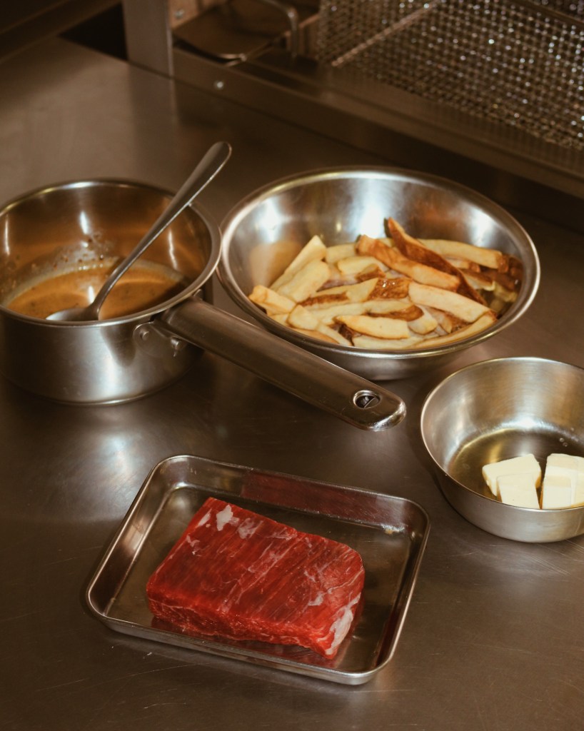 Close-up of a stainless steel kitchen countertop featuring a raw piece of red meat on a tray, two pots with contents, a bowl of French fries, and a small dish with cubes of butter.