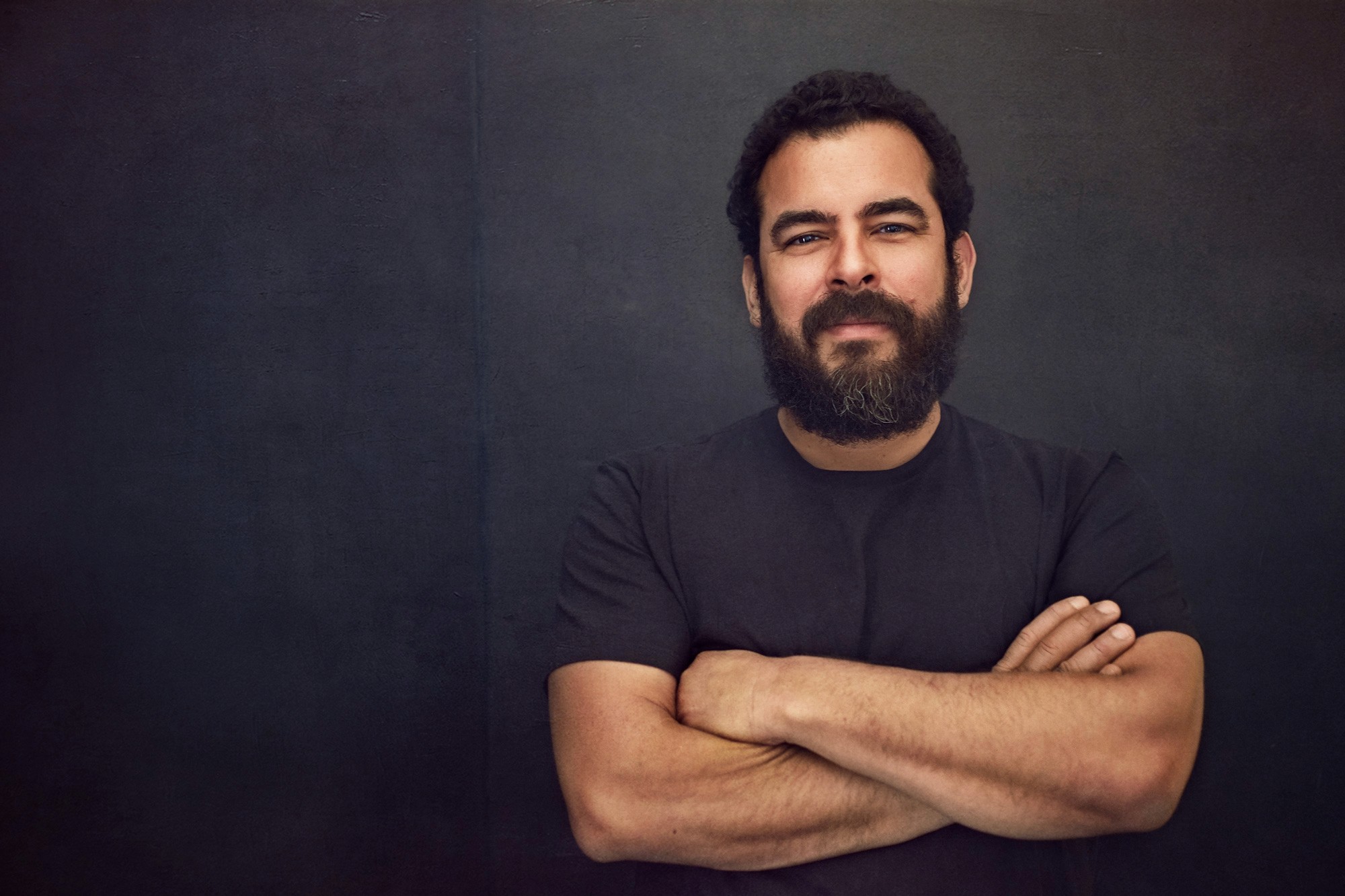 Studio headshot of bearded man in black t-shirt with arms crossed against dark textured background