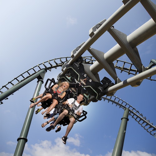 Twee mensen rijden op een omgekeerde achtbaan onder een zonnige, blauwe hemel met een paar wolken. Beiden lijken te genieten van de rit.