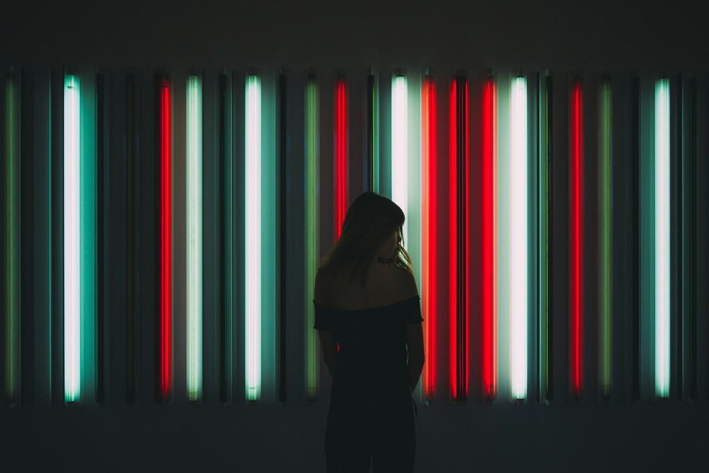 A silhouetted woman stands in front of a vibrant art installation featuring vertical neon lights in red, green, and white, creating a striking visual contrast.