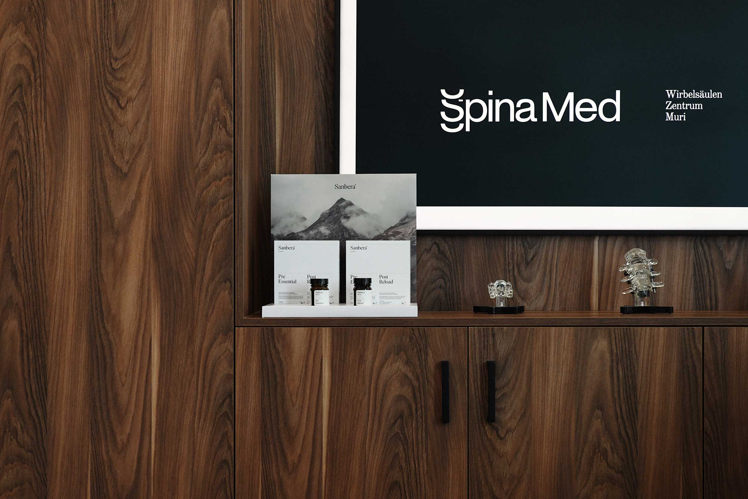 Holzvertäfelung mit Sideboard in der Praxis SpinaMed Wirbelsäulen Zentrum Muri, darauf Infomaterial und kleine Wirbelsäulenmodelle vor einem Bildschirm mit Praxislogo.