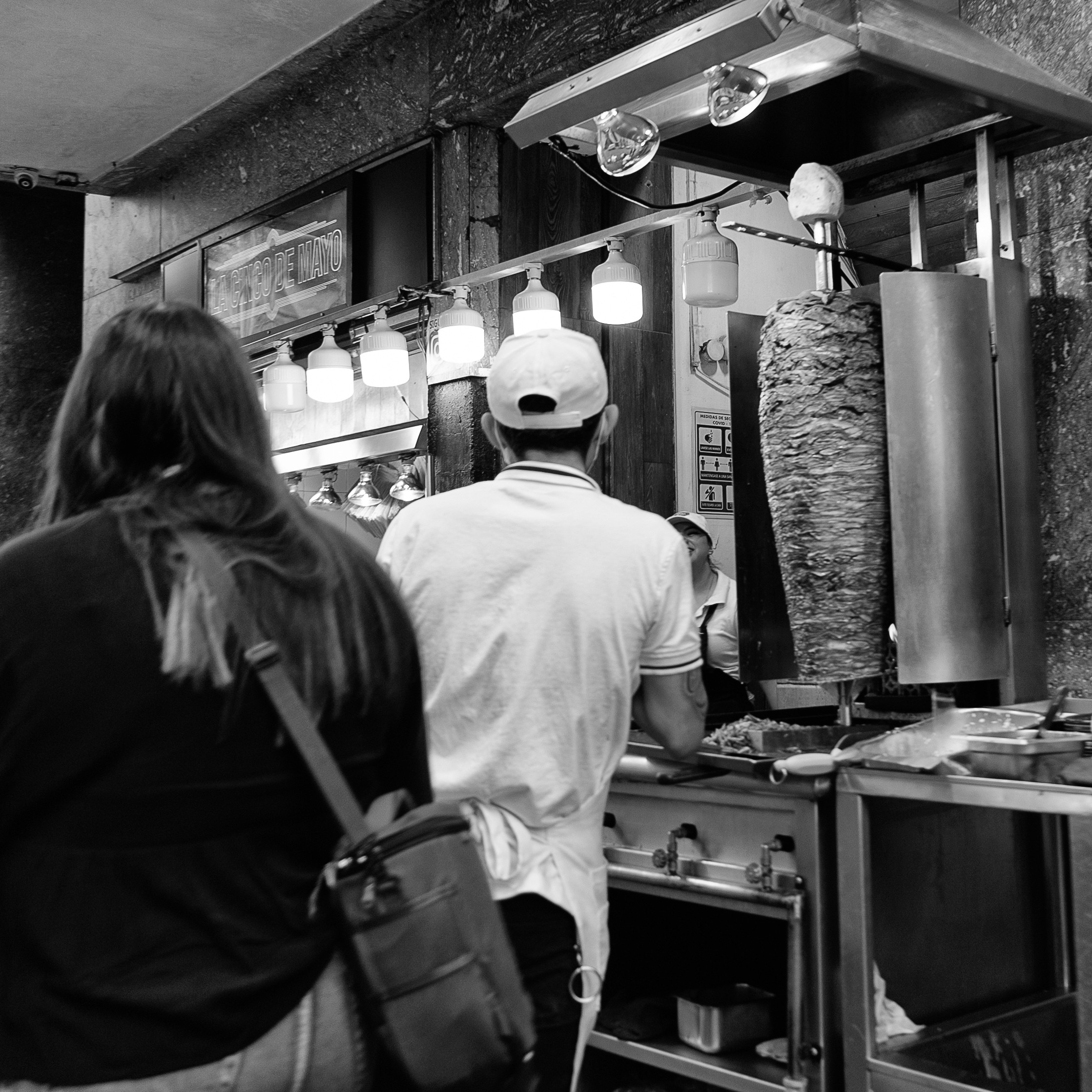 Black and white photo of a taco stand with a trompo of pastor meat and pineapple, a taquero at the grill, and customers waiting to be served.